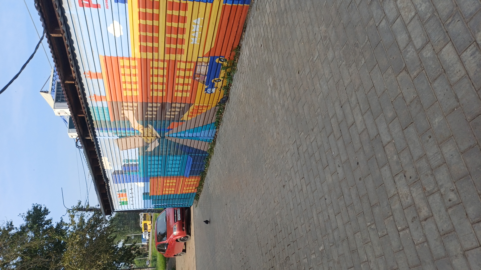 The image captures a street scene in Izhevsk, Russia, on a clear day, likely during daylight hours given the bright sunlight and clear blue sky. In the foreground, a textured grey brick-paved sidewalk or pedestrian area dominates the lower portion of the frame.
To the left of the sidewalk, a red car, possibly a sedan, is parked. The car's license plate is partially visible and reads "M931AK 18". A lone pigeon stands on the pavement in front of the car.
The background features a long, corrugated metal structure adorned with a vibrant mural. The mural depicts a stylized cityscape with buildings rendered in bold colors like blue, orange, and yellow. A small blue car is painted on the right side of the mural, and the word "MAMA PIZZA" is visible in white text above it. Above the corrugated structure, several air conditioning units are mounted, and a small section of a white awning or roof is visible. Further in the distance, a yellow bus and trees line a road, suggesting a typical urban or suburban environment. The overall impression is one of a commercial or service area with a decorative element on the building.