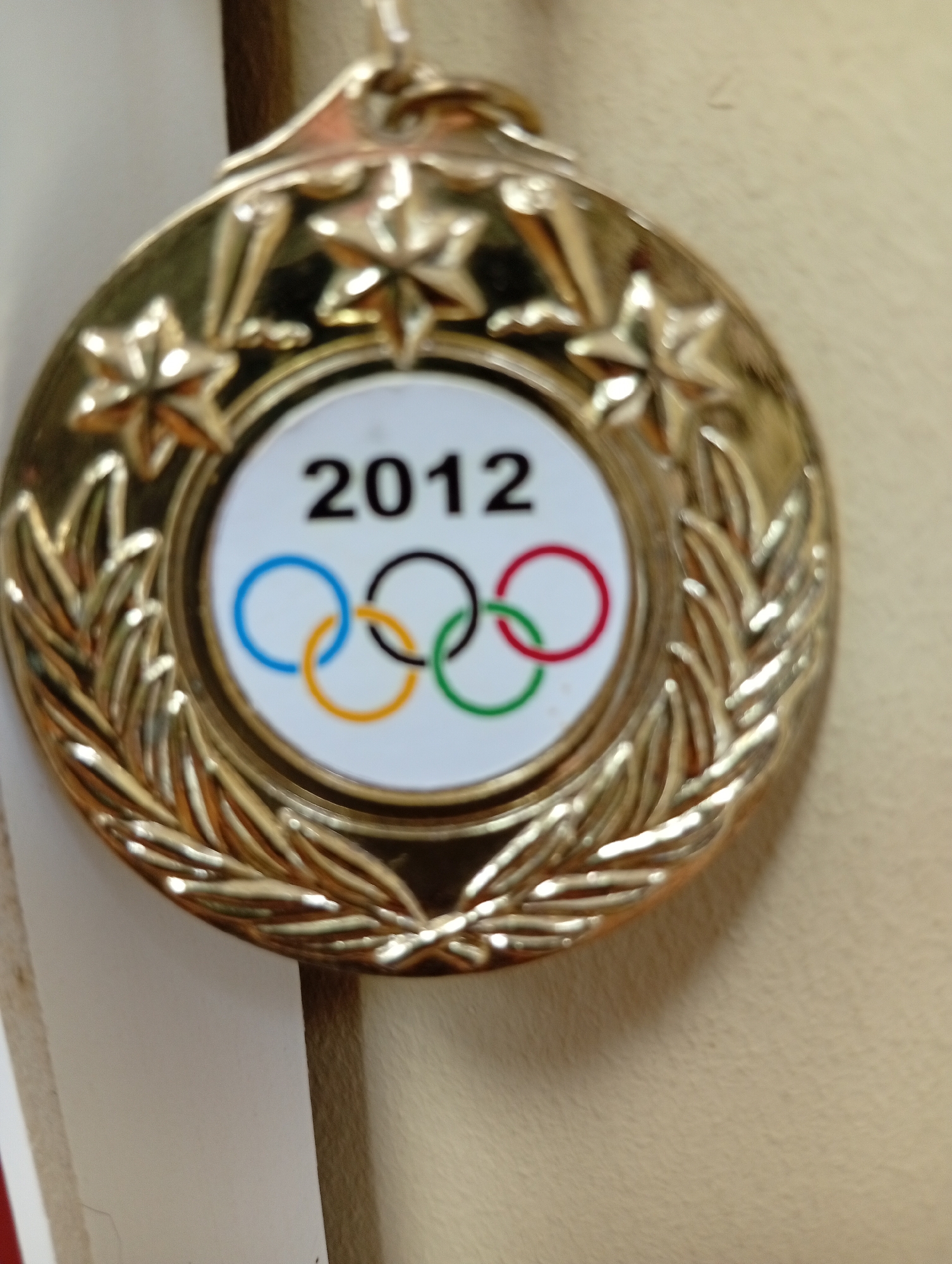 A close-up shot shows a gold Olympic medal from the 2012 games. The medal has stars embossed on its outer rim, and a laurel wreath design encircling the central emblem. The center features the Olympic rings in their traditional colors: blue, yellow, black, green, and red. The year "2012" is prominently displayed above the rings. The medal is suspended by a cord, which is partially visible at the top of the frame. The background is a plain, light-colored surface, possibly a wall or fabric. The location is not specified in the image.