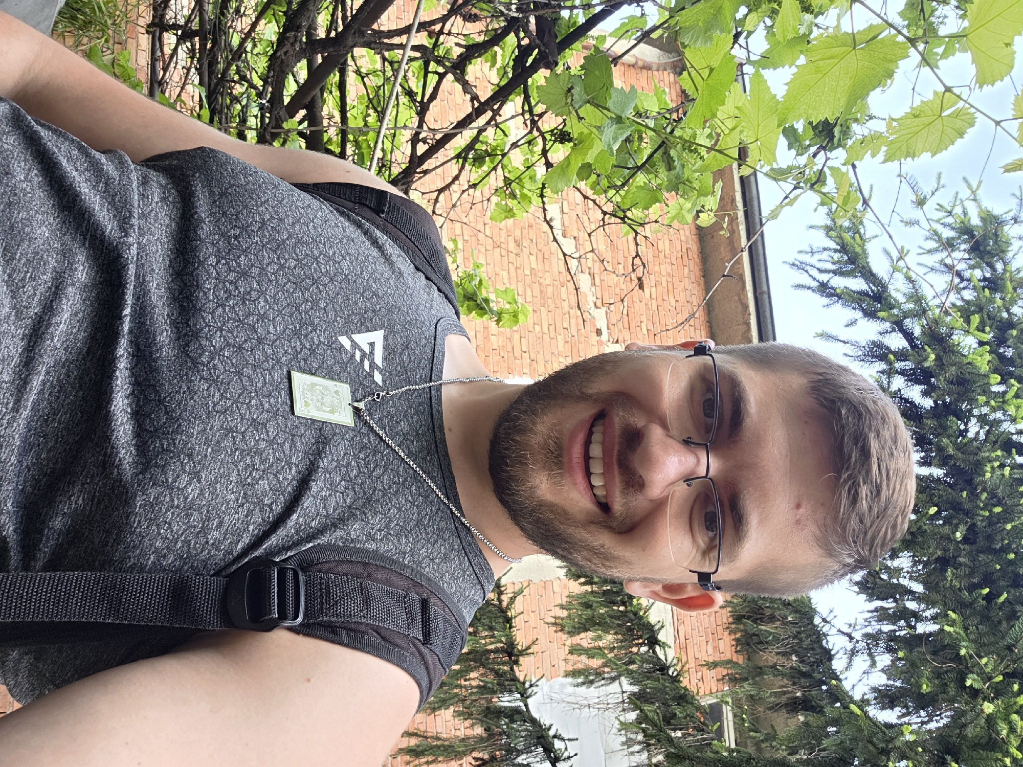 A medium shot depicts a smiling man with glasses and a beard, wearing a gray patterned tank top with a black backpack. He has a silver chain necklace with a rectangular pendant. The background features a brick building partially obscured by green foliage, including grape vines and an evergreen tree. The location is likely Yagodovo, Bulgaria based on the request context.