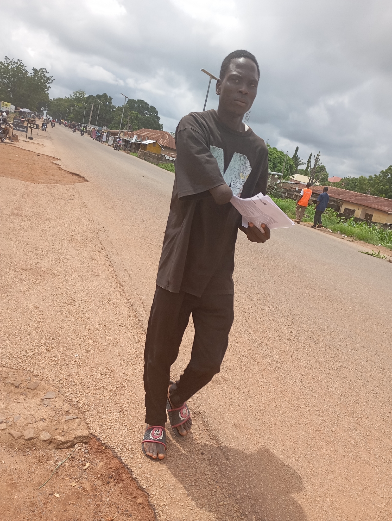 A full-body, low-angle shot shows a young Black man standing on a paved road in what appears to be a semi-urban area of Shendam, Nigeria, on a cloudy day. He is wearing a dark t-shirt with a graphic print and dark trousers, along with sandals. The man is holding several sheets of white paper in his right hand and looking slightly to his right. His left arm is bent at the elbow, with his hand resting on his hip.
The road is a light brown asphalt with some rough patches and cracks visible. To the left of the road, there is a dirt shoulder with some sparse vegetation. In the background, there are several buildings with corrugated metal roofs, some trees, and utility poles with streetlights. A few other people and motorcycles are visible further down the road, suggesting a populated area. One person in the background is wearing an orange shirt, and another is in a blue shirt. The sky is overcast with heavy gray clouds. The overall impression is of a bustling, everyday scene in a Nigerian town.