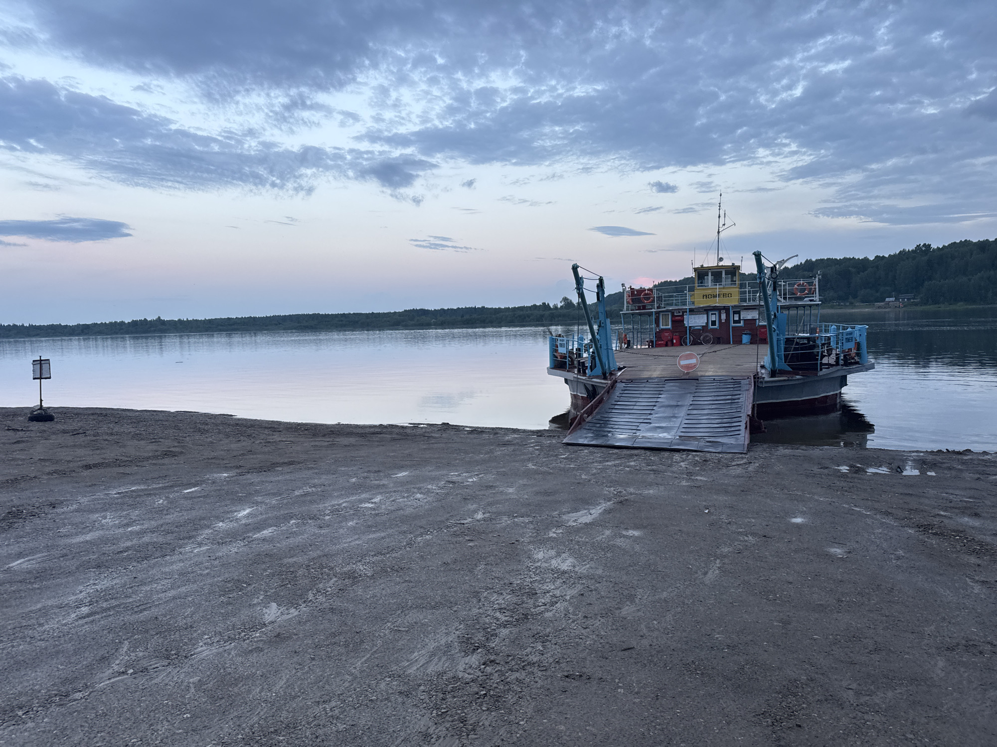 This image depicts a ferry docked at a shoreline on the Volga River near Tyulkino, Russia, during twilight. The sky displays a gradient of pale blue and pink hues, with scattered clouds. A ferryboat, painted with red and blue accents and a yellow superstructure, is positioned on the right side of the frame, its ramp extended to the shore. The ferry appears to be empty. The shore itself is a muddy, gravelly area with some scattered debris. In the distance, a dense forest lines the opposite bank of the river. A signpost with a white square on a pole stands on the left side of the shore. The water is calm and reflects the sky.