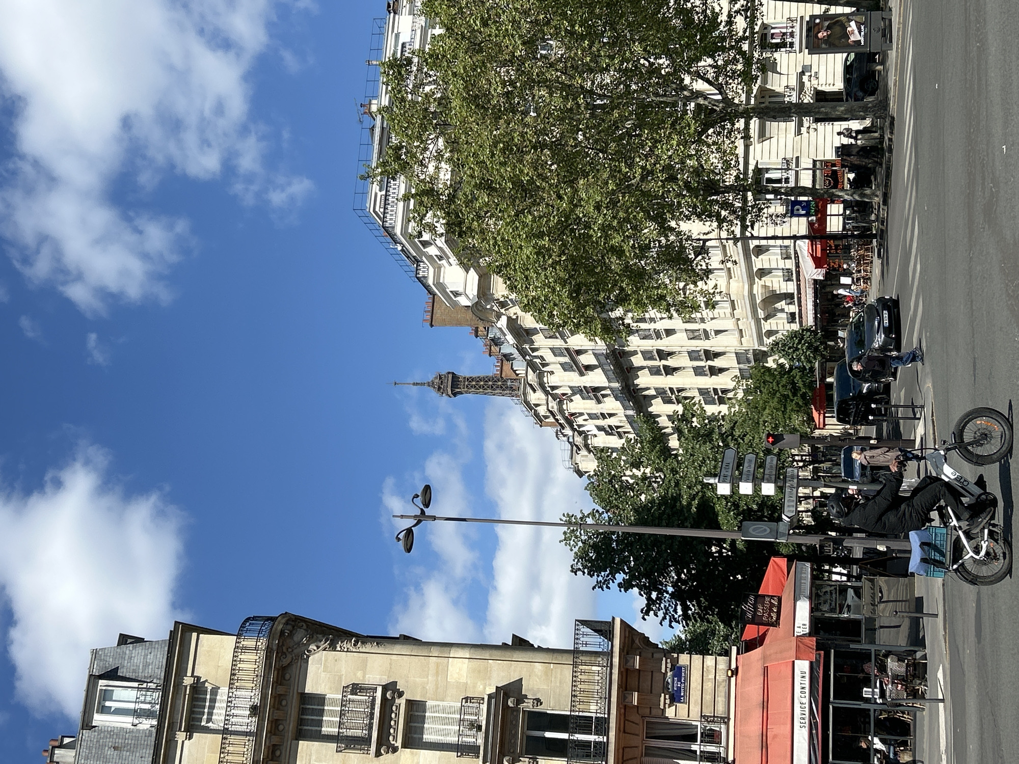 The image depicts a bright, daytime street scene in Paris, identified by the visible Eiffel Tower in the distance. The sky is a clear, vibrant blue with scattered white, fluffy clouds.
In the foreground, a classic Parisian building with ornate balconies and a grey slate roof is visible on the left. A tall, dark street lamp stands before it. The main street, likely paved, is busy with parked cars and features more traditional Parisian architecture, characterized by light-colored, multi-story buildings with numerous windows. Mature green trees with full foliage line the street, suggesting a spring or summer setting.
On the right side of the street, a person wearing dark clothing and a helmet is riding an electric bicycle, which has a blue crate attached to its back. The brand "APO" is visible on the bike frame. Nearby, a red traffic light and a pole with multiple white directional street signs are visible. An outdoor cafe with red awnings and seating is also present, with the text "SERVICE CONTINU" clearly readable on one awning. A partial sign for "PARKING 24/24" is also visible. A blue street sign at the bottom left distinctly reads "AVENUE LA MOTTE-PICQUET".
The upper section of the Eiffel Tower stands prominently in the background, peeking above the buildings and trees, confirming the location as the French capital. The scene conveys a sense of everyday urban life in Paris under pleasant weather conditions.