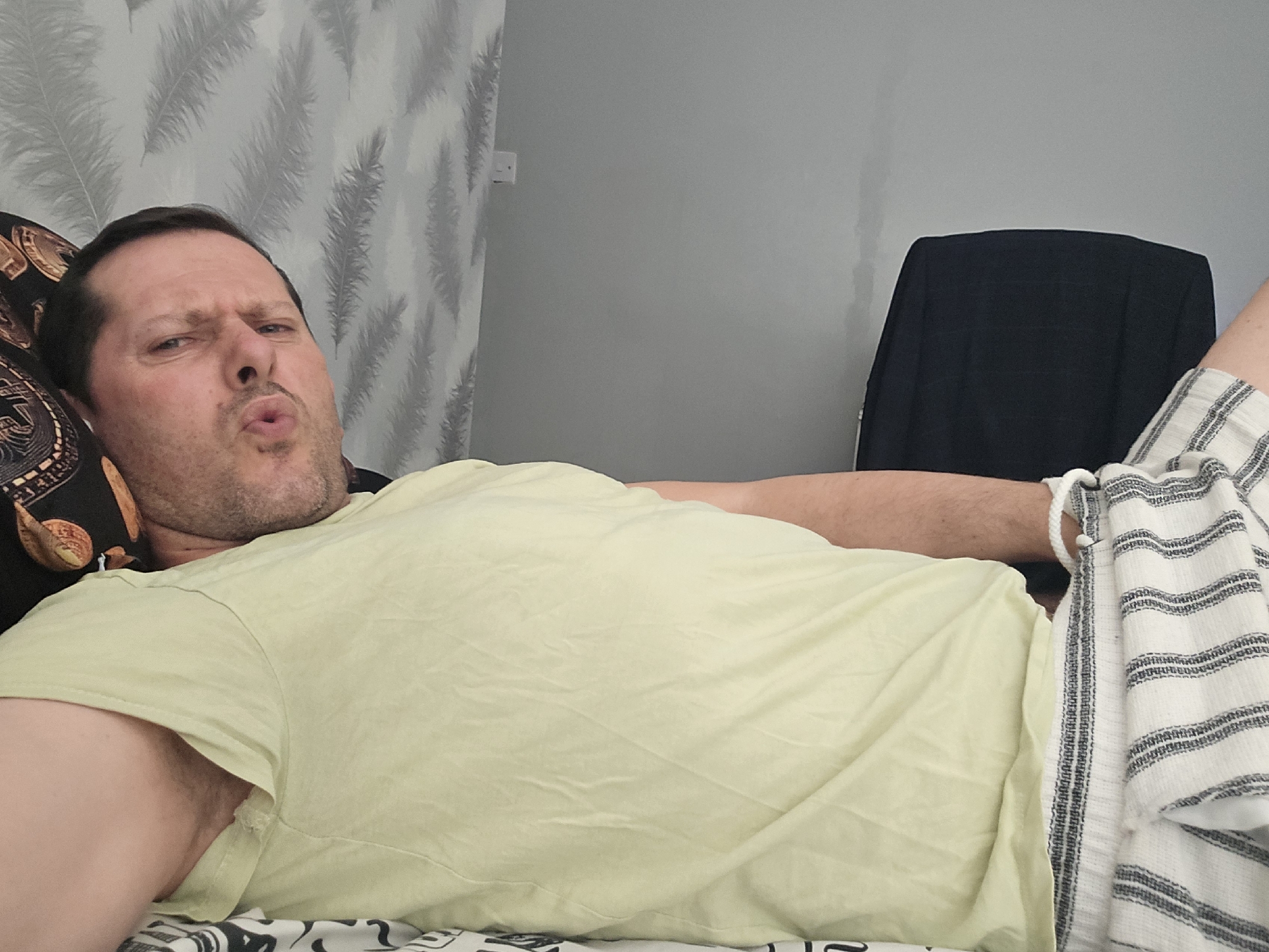 A Caucasian male lies on a bed with a patterned duvet. He wears a pale yellow t-shirt and striped shorts. His head rests on a pillow with a dark, ornate design. The background features a wall with a leaf pattern and a solid grey wall with a black chair. The male is making a pouting face. The location cannot be determined from the image.