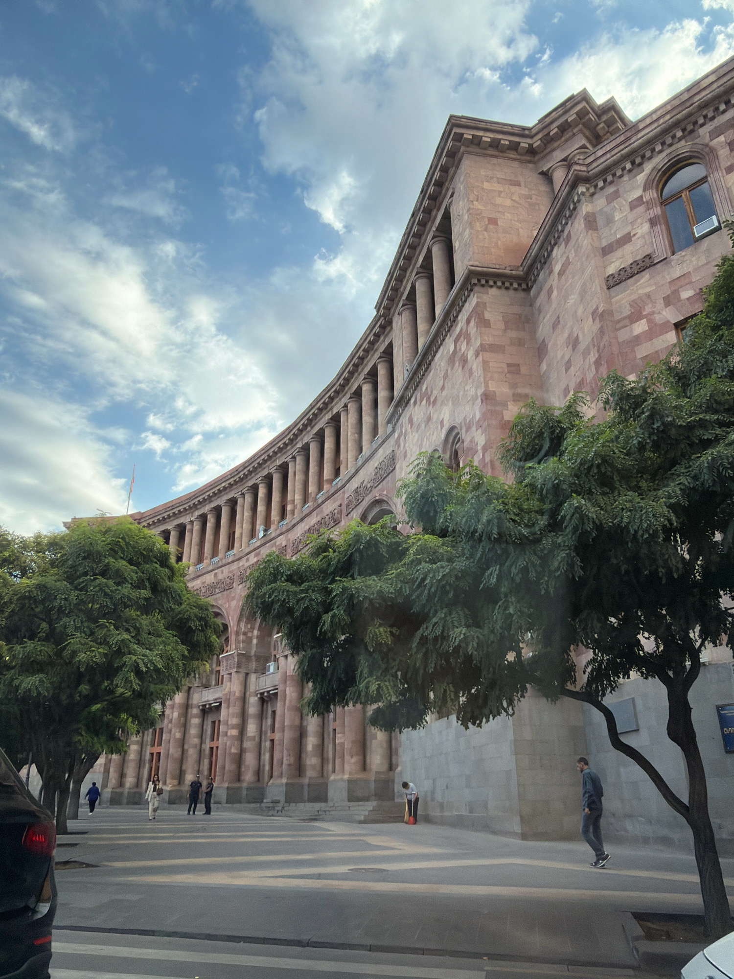The image shows a wide, curved building with classical architecture in Yerevan, Armenia. The building is made of pinkish-brown stone and features a colonnade along its facade. Tall trees with lush green foliage are planted in front of the building, partially obscuring the lower levels. The sky is partly cloudy, with patches of blue sky visible. Several people are seen walking on the sidewalk in front of the building. One person in a white suit walks towards the viewer, accompanied by two individuals in dark uniforms. Another person in a blue jacket walks away from the viewer. Further to the right, a person is sweeping with a red broom and a red bucket. A man in a dark shirt and pants walks towards the right, past a tree. The ground in front of the building is paved with stone slabs and there is a pedestrian crossing marked on the road in the foreground. A black car is partially visible on the left side of the image. The building is likely a government building or a significant public institution, given its grand design and prominent location.