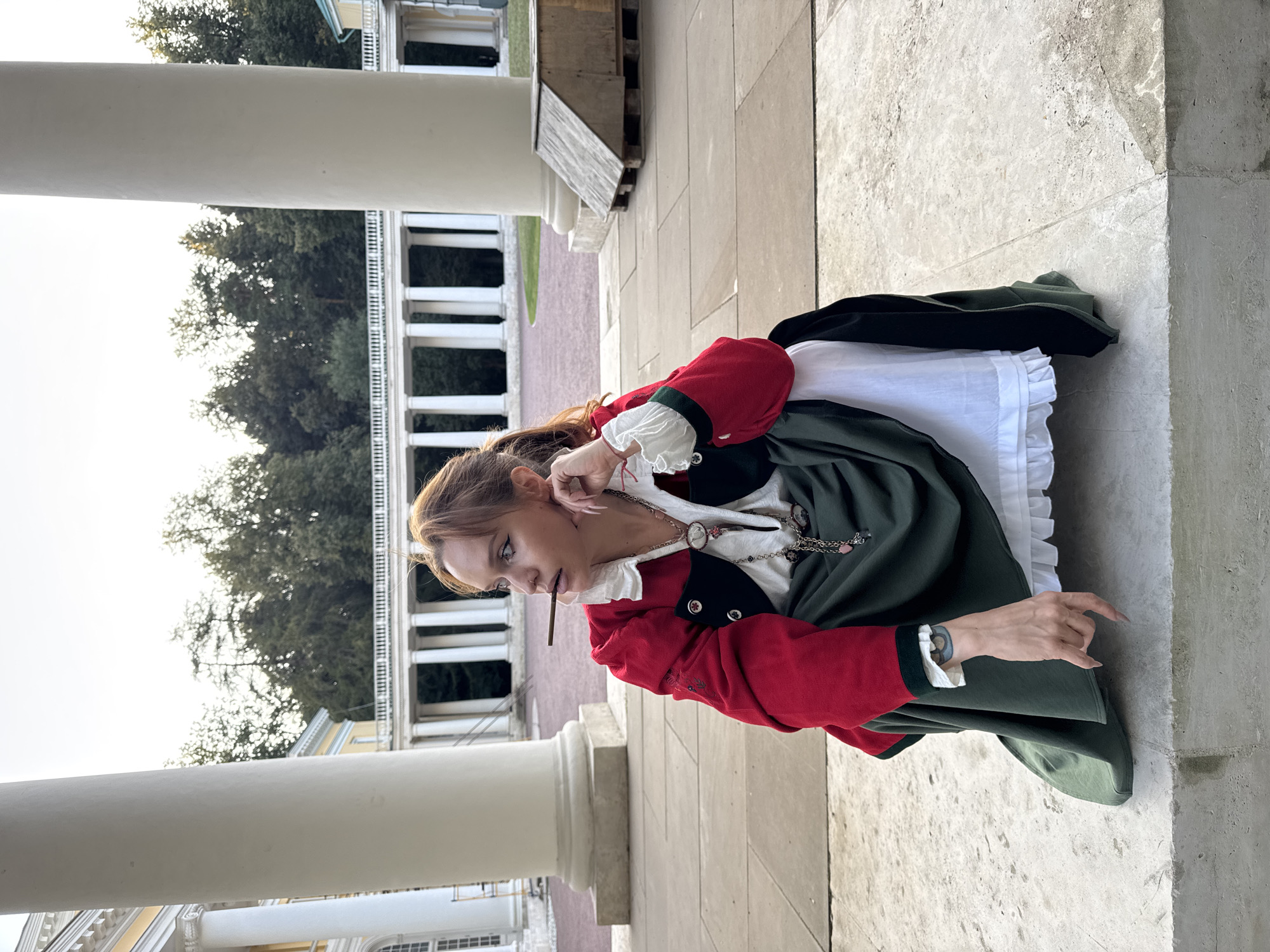 A young woman with auburn hair pulled back in a ponytail is squatting on a stone ledge in front of a neoclassical colonnade in Arkhangel'skoe, Russia. She is wearing a historical costume consisting of a red jacket with gold trim and black accents, a white ruffled blouse, a dark green skirt, and a white underskirt. She has a dark object, possibly a cigar or pencil, in her mouth. Her left hand rests on her cheek, and her right hand is extended downwards, touching the stone. The background features a long, white colonnade with a light-colored building behind it, and trees further in the distance. The lighting suggests it is late afternoon or early evening.