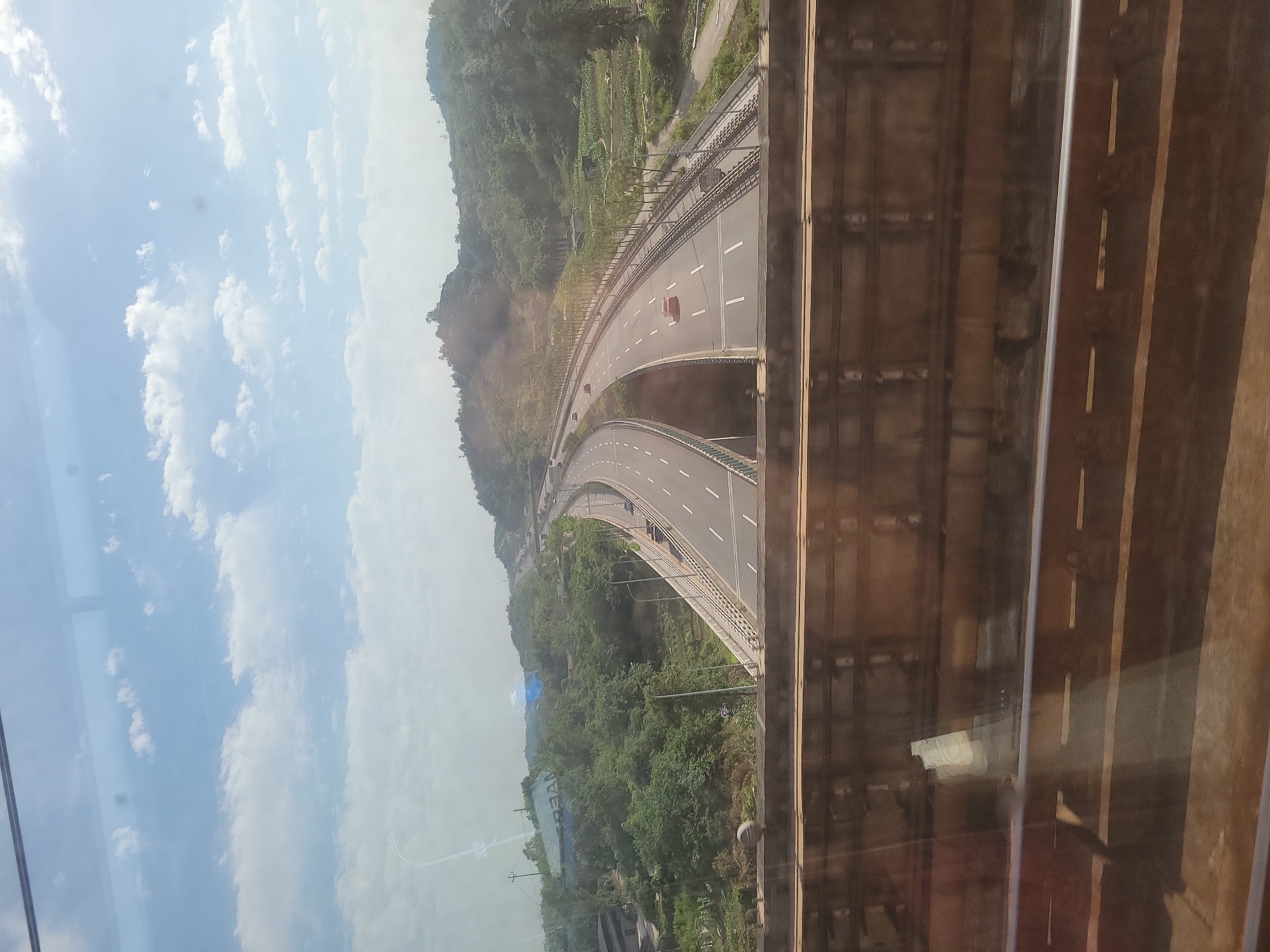 The image shows a view of a highway curving through a mountainous, forested area, as seen from what appears to be a high vantage point, possibly through a window. The highway has multiple lanes and some traffic. The sky is visible with clouds. The location is near Xiema Subdistrict, China.