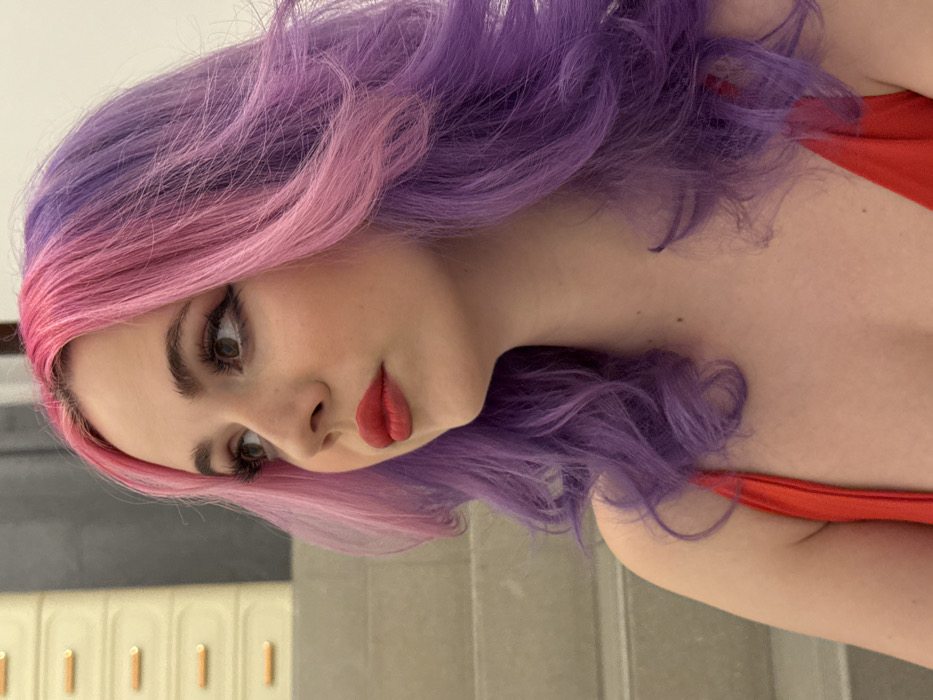 A woman with striking pink and purple hair, styled in loose waves, is the focal point. She's wearing red lipstick and a red top. Her gaze is directed downwards with her head tilted slightly. The background appears to be a bathroom with light-colored tiling and ornate detailing. The overall perspective is from a slightly elevated angle.