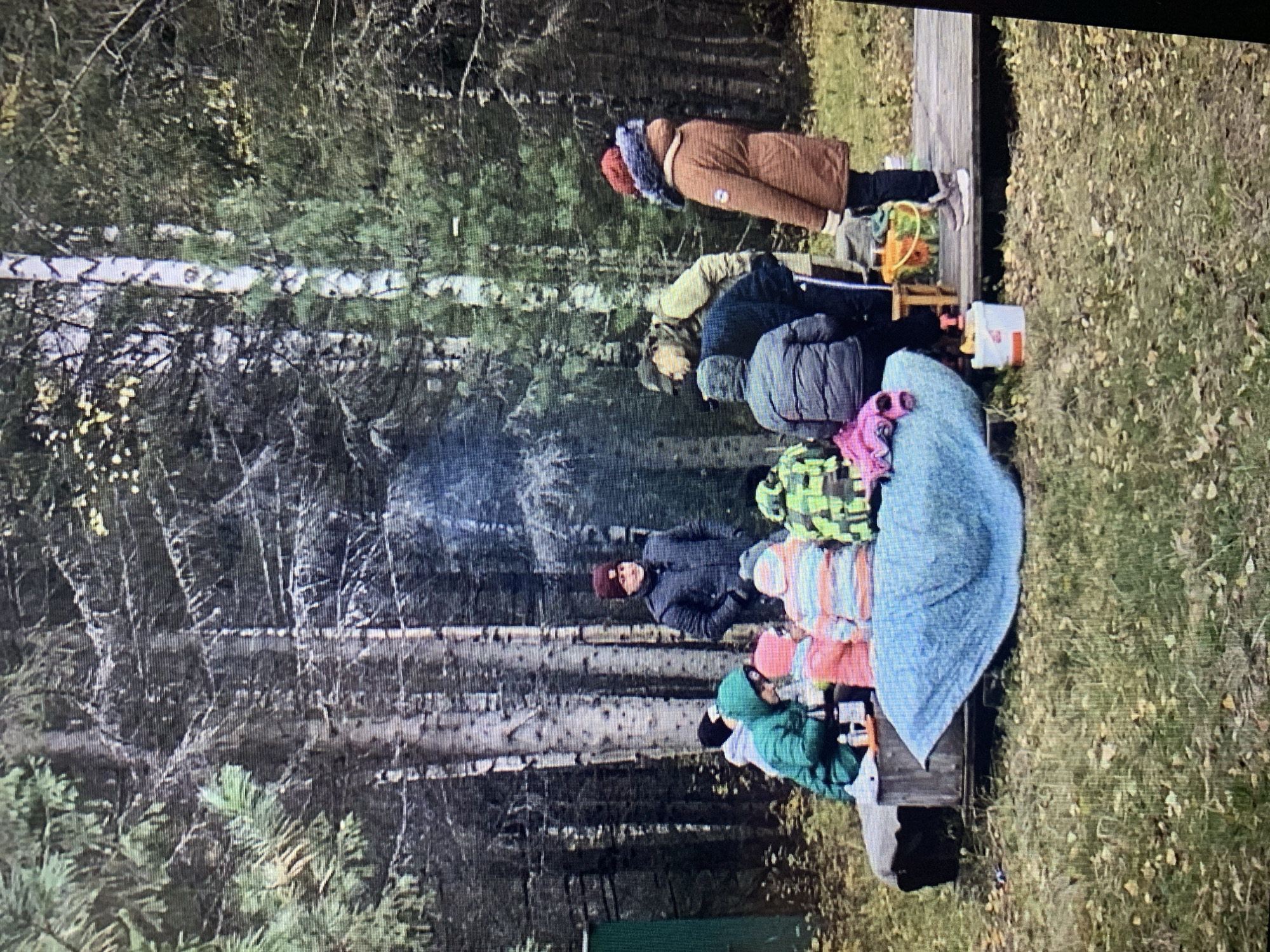 A group of people are outdoors, possibly in a park or wooded area, near Izhevsk, Russia. Trees are visible. Several individuals are sitting or standing around a low structure, likely a bench or platform. People wear winter clothes like hats and coats. There's a fire with smoke rising. Some objects, possibly toys or containers, are on the platform. The ground is covered with grass and fallen leaves. The setting seems to be a recreational area or an outdoor gathering place.