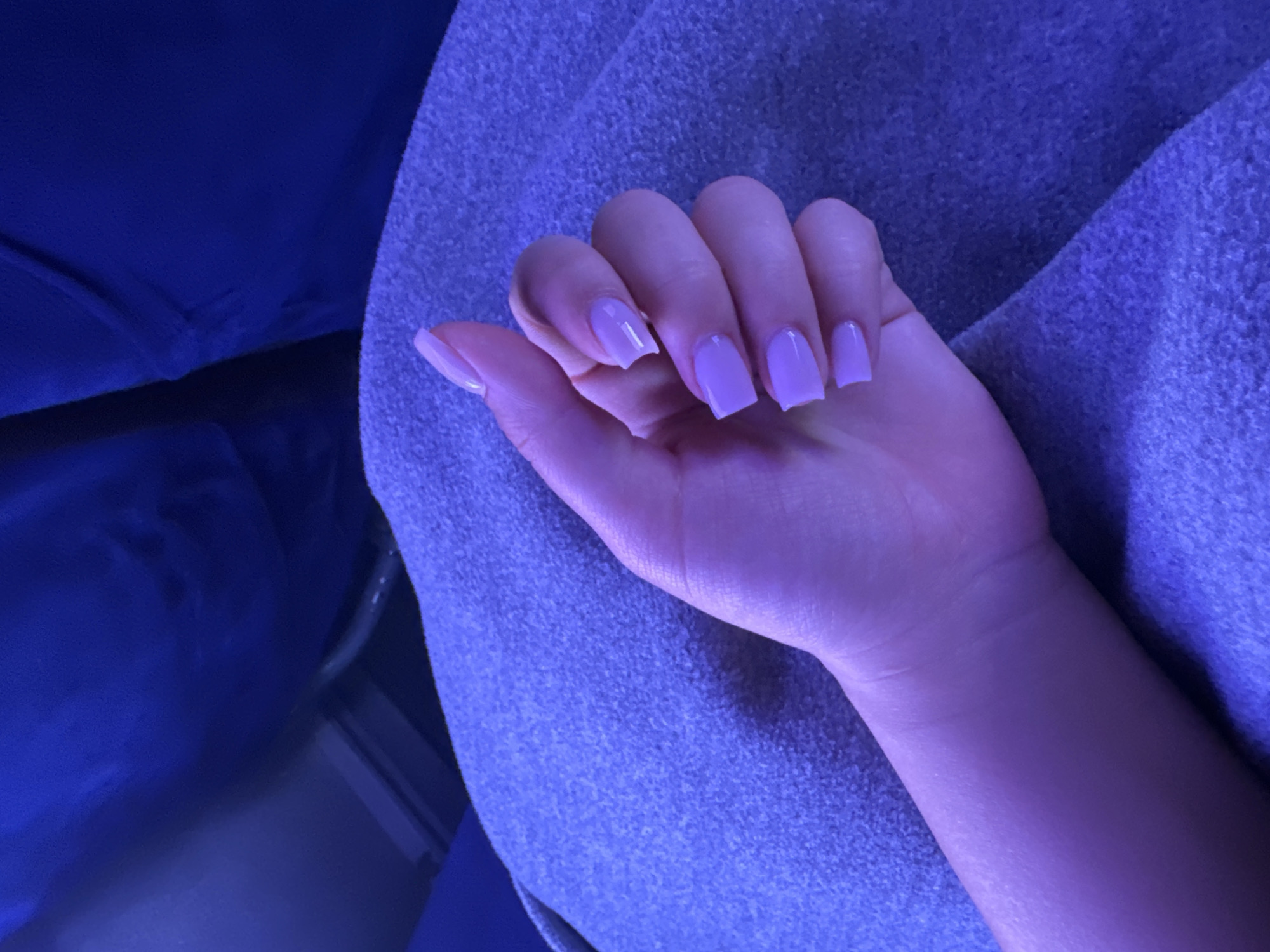 A close-up, overhead shot shows a person's left hand with neatly manicured square-shaped nails painted in a light lavender color, resting on a textured, light purple blanket. The fingers are slightly curled and relaxed. In the background, out of focus, is a dark blue textured surface, possibly a seat or fabric. The lighting in the image is predominantly a cool, blue-purple hue, suggesting artificial lighting. The setting appears to be indoors, with no visible indication of a specific location in a city.