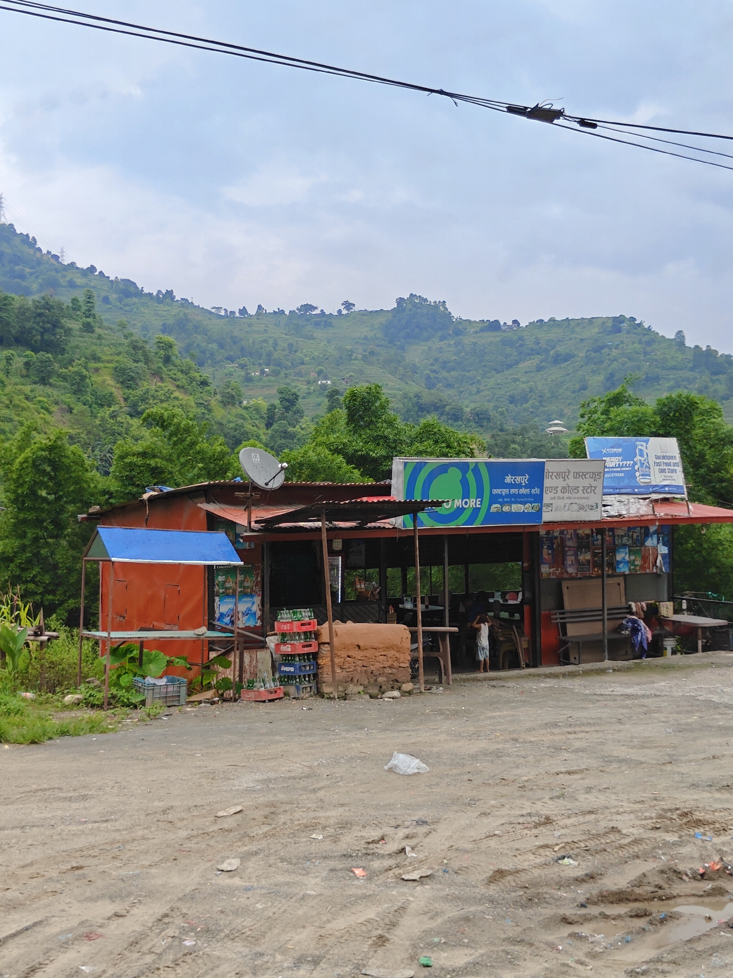 The image shows a roadside shop in Thakre, Nepal, with a green, terraced hillside in the background under a cloudy sky. The shop has a red tin roof supported by wooden poles, and an orange metal structure with a blue tin roof to the left. A satellite dish is mounted on the main roof. Signs above the shop display advertisements in Nepali and English, including "MORE" and "Gorakhpur Fast Food and Cold Store." Bottles of Coca-Cola and Pepsi are stacked outside the shop. A small dirt road with tire tracks and scattered debris is in the foreground. A young boy in a white shirt and shorts stands near the entrance of the shop. The environment appears to be rural, with dense vegetation on the hills.