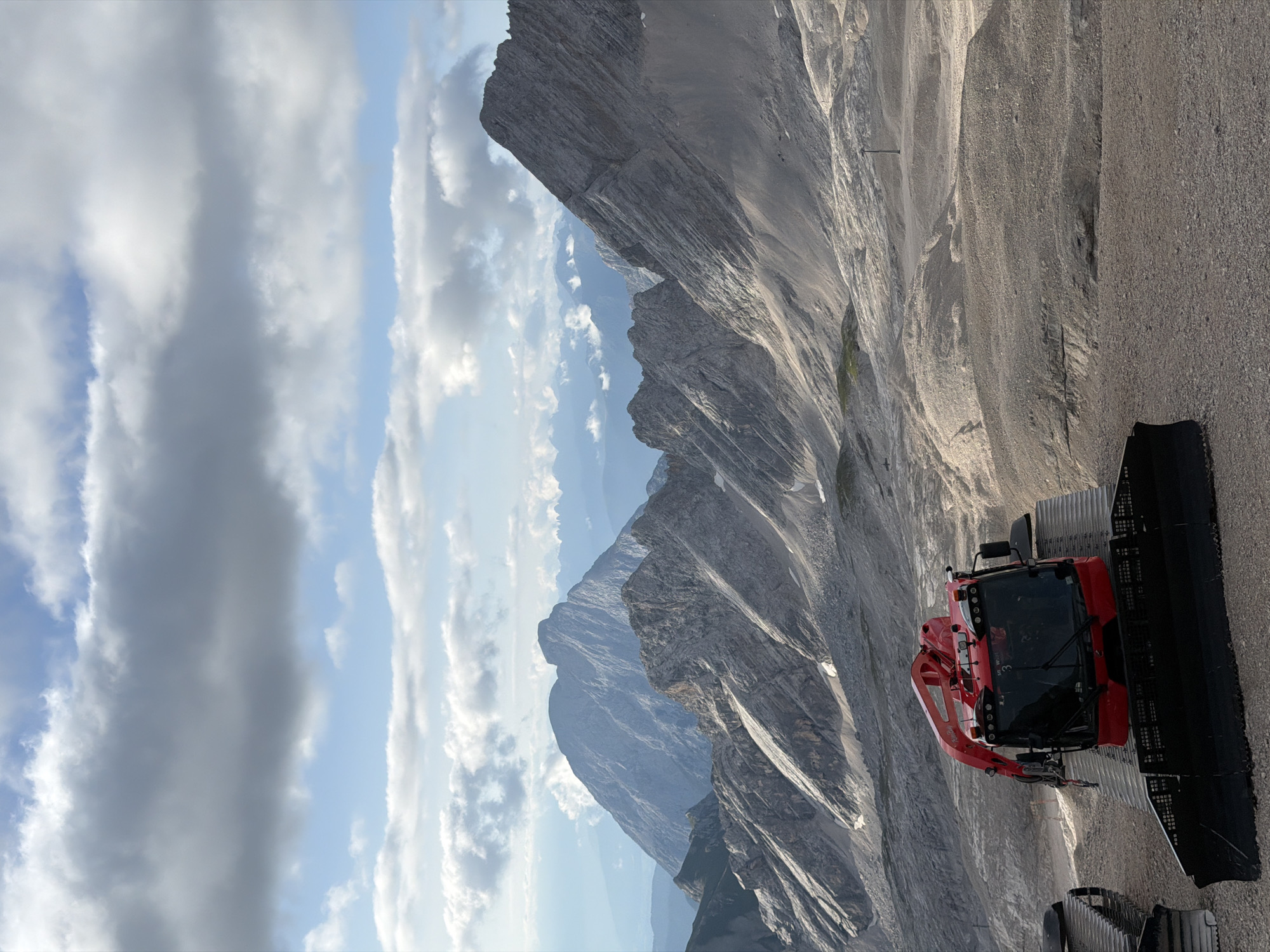 The image depicts a scenic mountainous landscape, likely in the Bavarian Alps near Garmisch-Partenkirchen, Germany. In the foreground, a bright red snow groomer, a vehicle used for maintaining snow surfaces, is parked on a gravelly terrain. The vehicle has a large, black plow in front and tracks for mobility. Behind the snow groomer, a vast expanse of rocky mountain slopes stretches into the distance, with layers of peaks fading into a hazy blue sky. The sky is partly cloudy, with a mix of white cumulus clouds and patches of blue. The overall impression is one of a rugged, high-altitude environment, possibly during a break in snowy conditions.