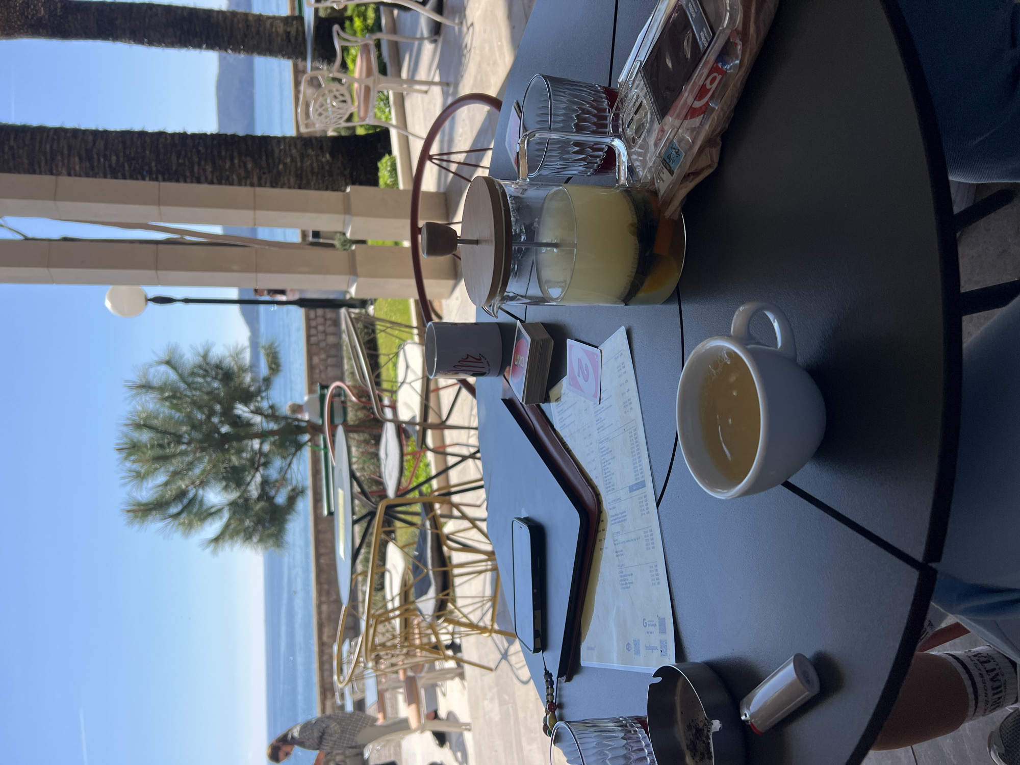 The scene is a table at an outdoor café in Rafailovići, Montenegro, overlooking the Adriatic Sea. The table holds a glass teapot of light yellow liquid with fruit pieces, a glass, a cup of tea, a menu, a smartphone, a package of sugar, an ashtray, and a lighter. A playing card is also visible. In the background, palm trees and other seating can be seen along the coastline, along with building columns and a streetlamp. The sea and sky are blue. A person wearing a grey shirt and black pants is near the table. Another person is seated near the table.