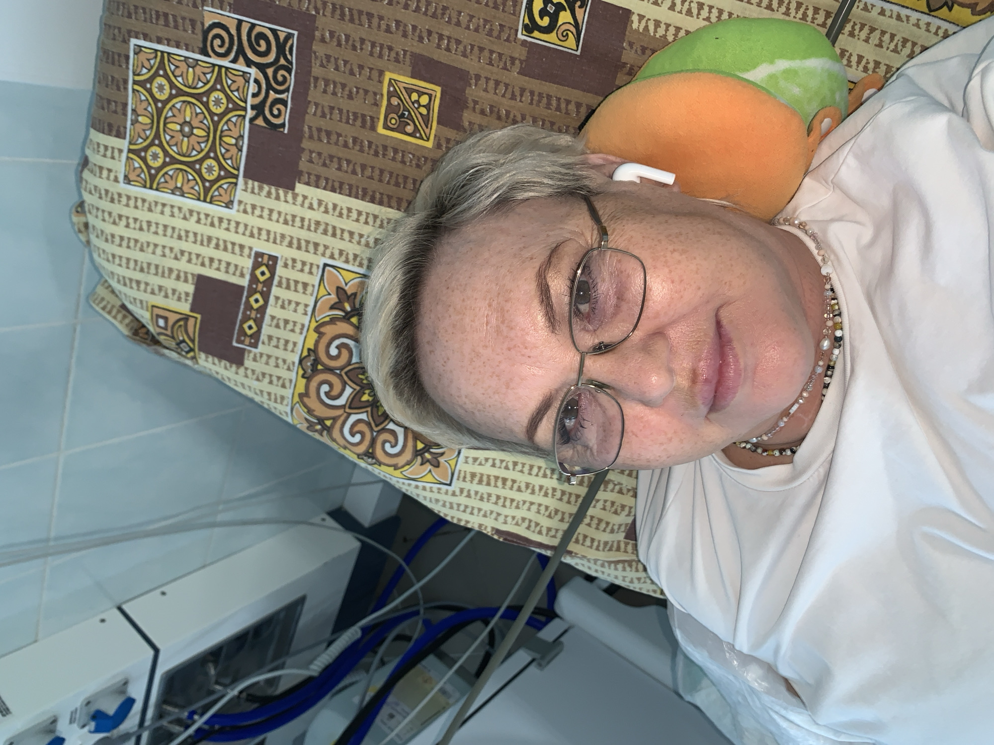 An image taken from an overhead perspective captures a middle-aged to older woman lying down, looking towards the camera. She has fair skin with numerous freckles, short blonde and grey hair, and wears wire-rimmed glasses. White wireless earbuds are visible in her ears, and she wears a multi-strand beaded necklace around her neck, complementing her simple white t-shirt.
She is resting on patterned bedding featuring a mix of brown, beige, yellow, and black geometric designs. A distinctive green and orange plush toy, resembling a fruit slice, is tucked behind her head, possibly serving as a small pillow or comfort item. In the background, to the lower left, light blue tiled walls are visible, along with specialized electronic or medical equipment. This equipment includes white casings, numerous blue cables, and various ports, suggesting a clinical or medical setting. The woman maintains a calm, neutral expression. The scene is located in Chaikovsky, Russia.