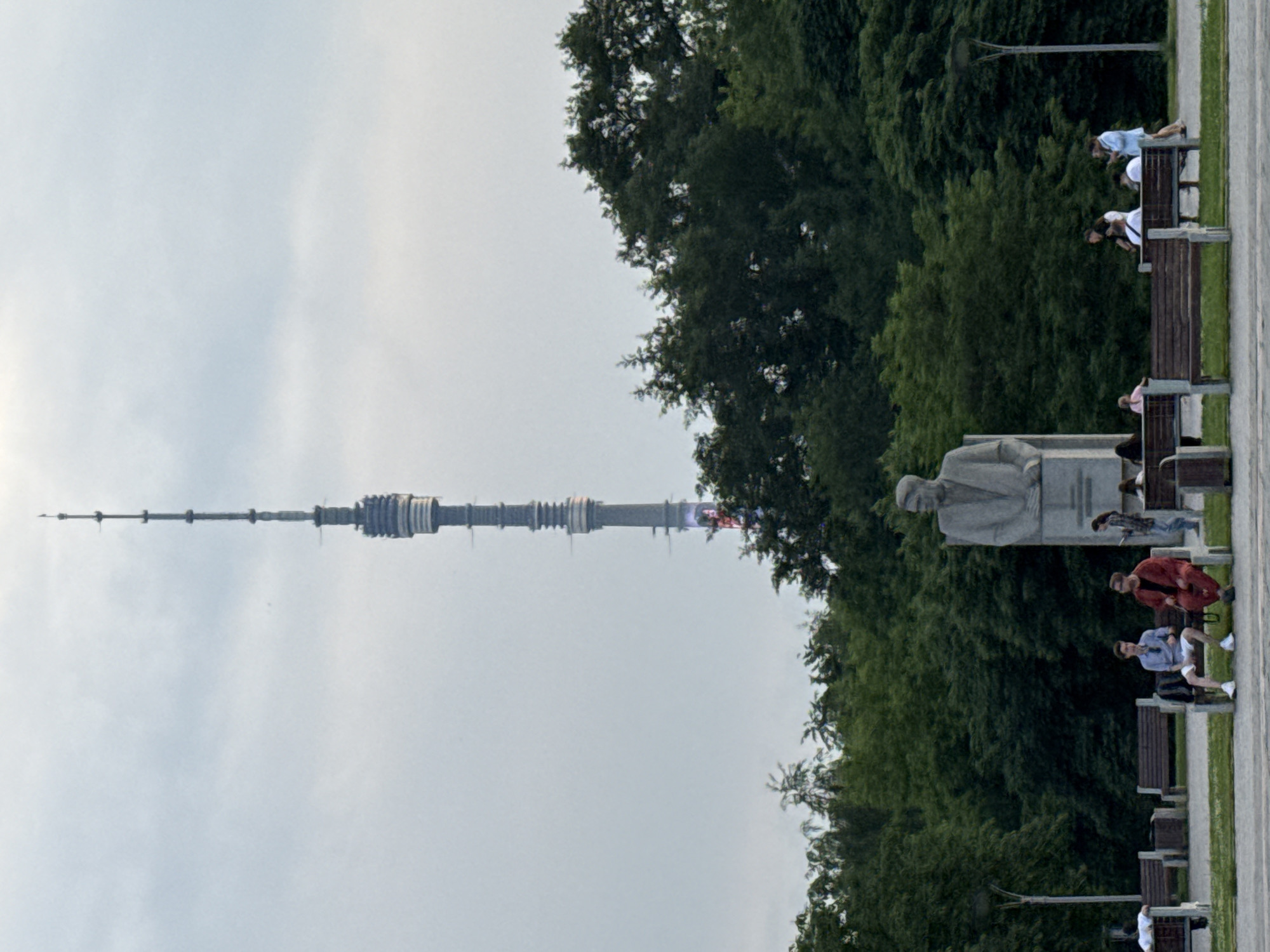 The image depicts a wide shot of a park area with a tall, slender telecommunications tower in the background. The tower, likely the Ostankino Tower, is a prominent landmark in Moscow, Russia. In the foreground, several people are seated on park benches, some interacting with each other. A large stone statue of a man in a suit is visible to the right of the frame. Lush green trees form a dense backdrop behind the statue and park seating area. The sky is overcast, suggesting cloudy weather.