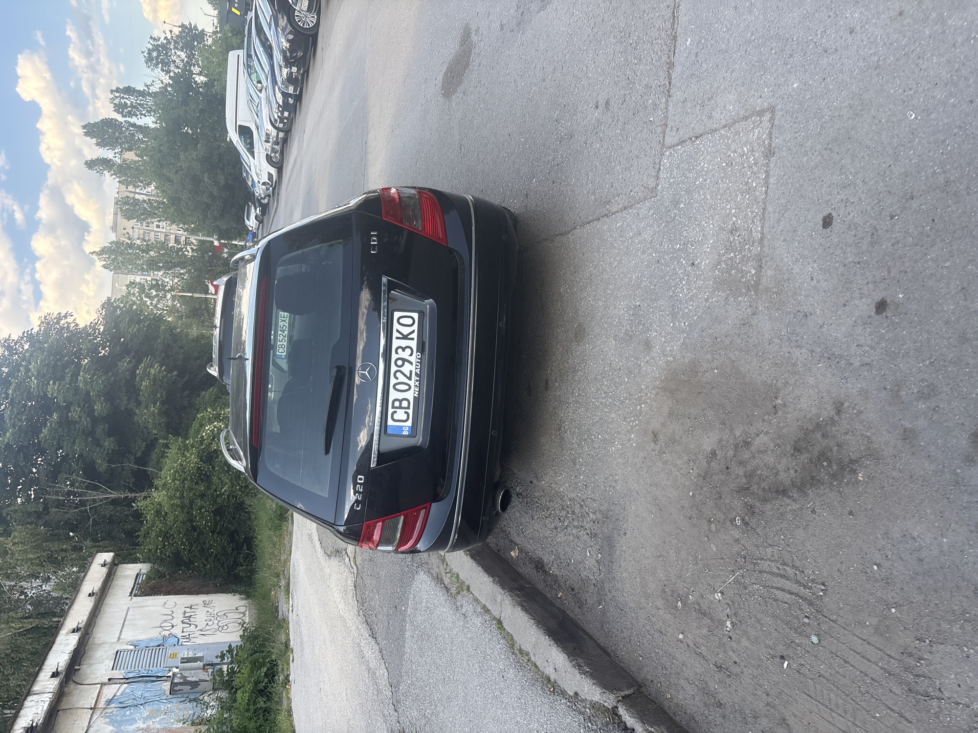 In Sofia, Bulgaria, on a sunny day, a black Mercedes-Benz C220 CDI wagon is parked on the right side of an asphalt road, adjacent to a curb. The car's license plate reads "CB 0293 KO". In the background, apartment buildings and trees are visible. To the left of the car, a dilapidated building with graffiti on its wall stands. The street is lined with parked vehicles, including vans and other cars. The sky is partly cloudy.