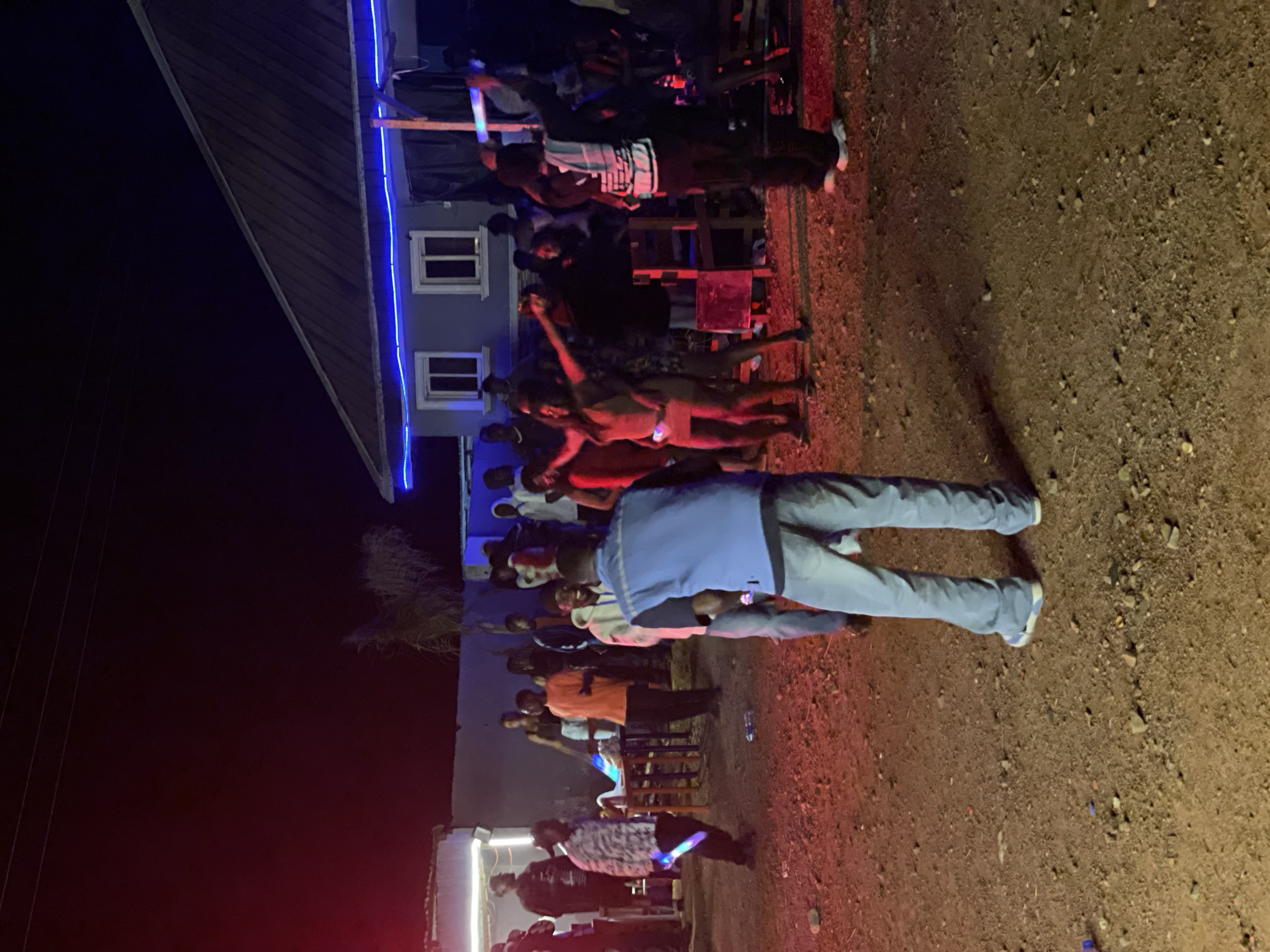 The image depicts an outdoor gathering at night in Durumi, Nigeria. A group of people are congregated in a dirt-covered area, illuminated by red and blue lights. In the background, a building with blue strip lights along its eaves is visible. Several individuals are dancing or socializing. Some are holding glowing objects that appear to be light sticks or similar party accessories. The ground is uneven and scattered with small rocks and debris. A few tables and chairs are present, suggesting a casual event or a makeshift outdoor venue. The overall atmosphere appears to be a lively party or social event taking place in a somewhat informal setting.