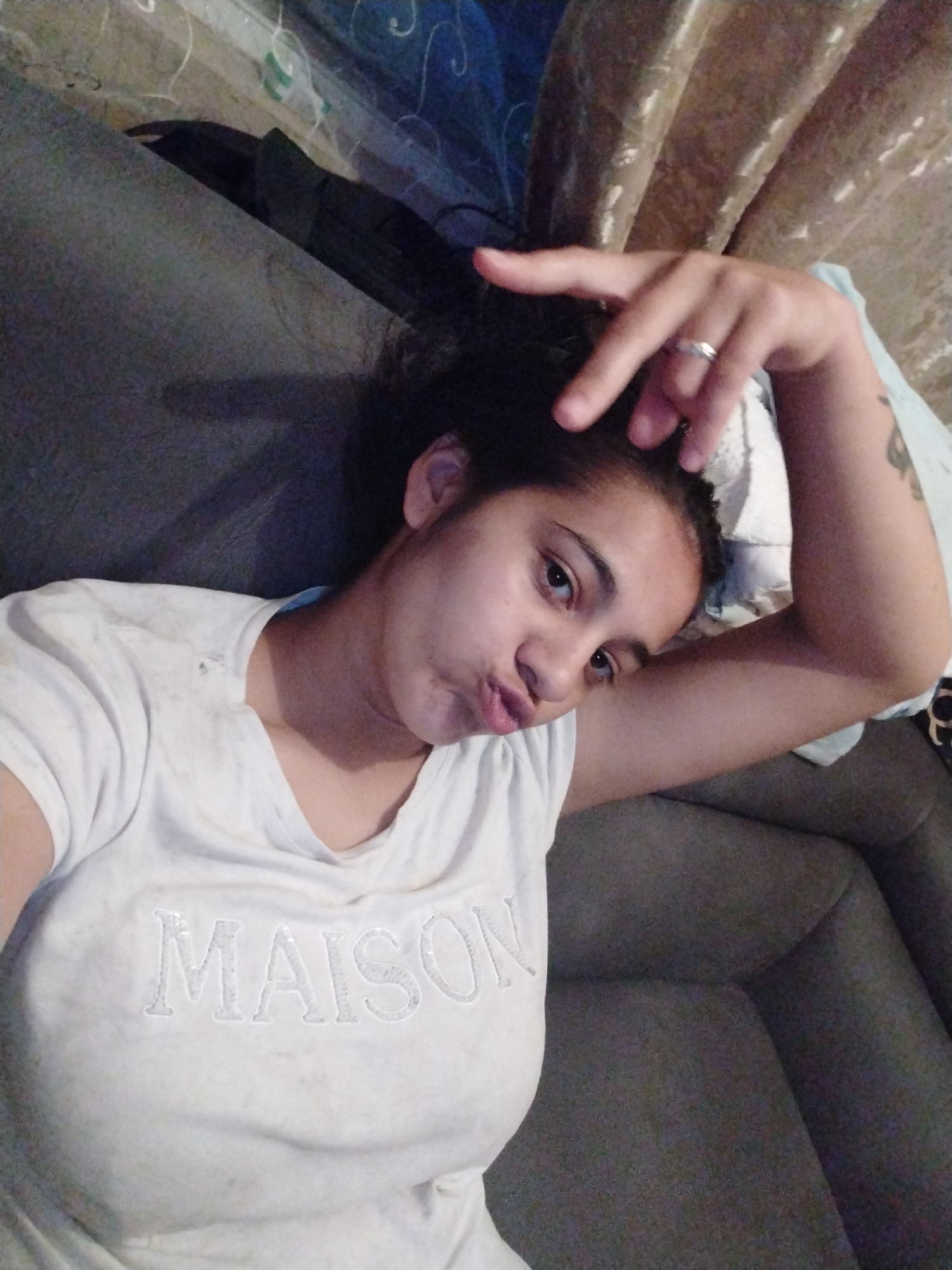 A person is lying on a gray couch and is wearing a white T-shirt with the word "MAISON" on it. The person has dark hair in a messy bun. They are making a peace sign with their right hand, and are wearing a ring on their ring finger. The location appears to be indoors, possibly a home. The city is Unknown, Unknown.