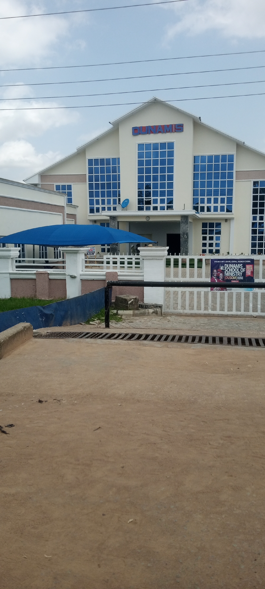 The image depicts the exterior of a building identified as the "DUNAMIS INTL. GOSPEL CENTRE, JALINGO CENTRAL". The building features a prominent "DUNAMIS" sign in red and blue letters above a facade with large, blue-tinted windows. A blue canopy shades an area in front of the building, supported by white pillars. A white fence runs along the front of the property, and a black metal railing is visible in the foreground. A poster for "DUNAMIS SCHOOL OF MINISTRY" is attached to the fence. The surrounding environment appears to be a street or a pathway with some grass and dirt. The sky is overcast with some clouds.