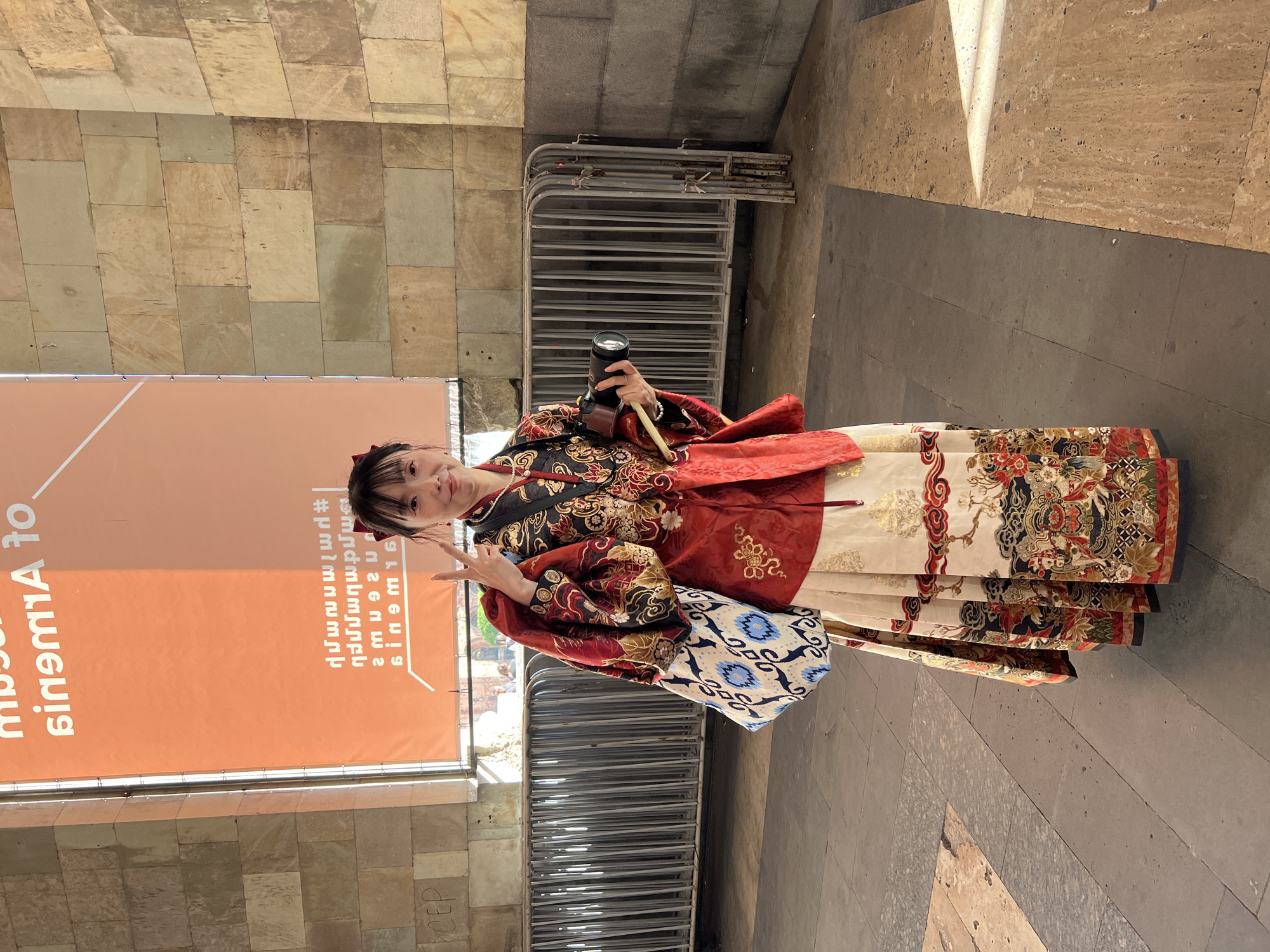 A woman stands in Yerevan, Armenia, wearing a red and gold ornate kimono and a blue and white patterned bag. She is holding a camera in her right hand and making a peace sign with her left. To her left, there is an orange banner with Armenian script. Behind her are silver metal barriers. The ground is paved with gray stone tiles and bordered by beige brick walls on both sides. The lighting suggests it is daytime.