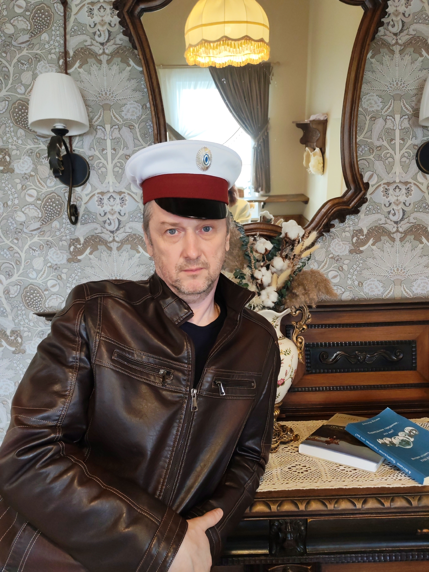 The image depicts a middle-aged man wearing a brown leather jacket and a peaked cap with a white top, red band, and black visor. The cap has a small emblem on the front. He is standing indoors, possibly in a room with antique-style furnishings and patterned wallpaper. Behind him is an ornate wooden frame, likely part of a mirror or dresser, adorned with a vase containing cotton branches. Two books are placed on the surface next to the vase. The setting may be in the city of Sarapul, Russia. The man is looking directly at the camera with a neutral expression.