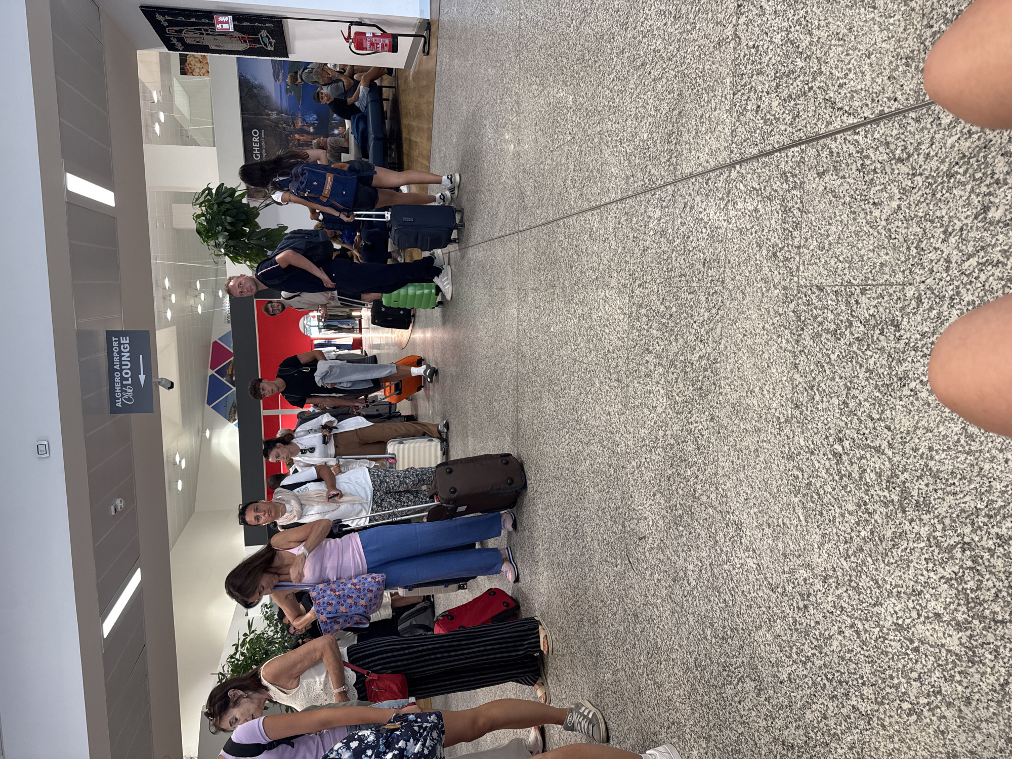 The image shows a queue of people in an airport terminal. The airport's name, Alghero Airport, is visible on a sign in the upper part of the image. Several individuals are standing in line, some with luggage. The floor is a speckled granite. The overall scene depicts a typical airport environment with passengers waiting.