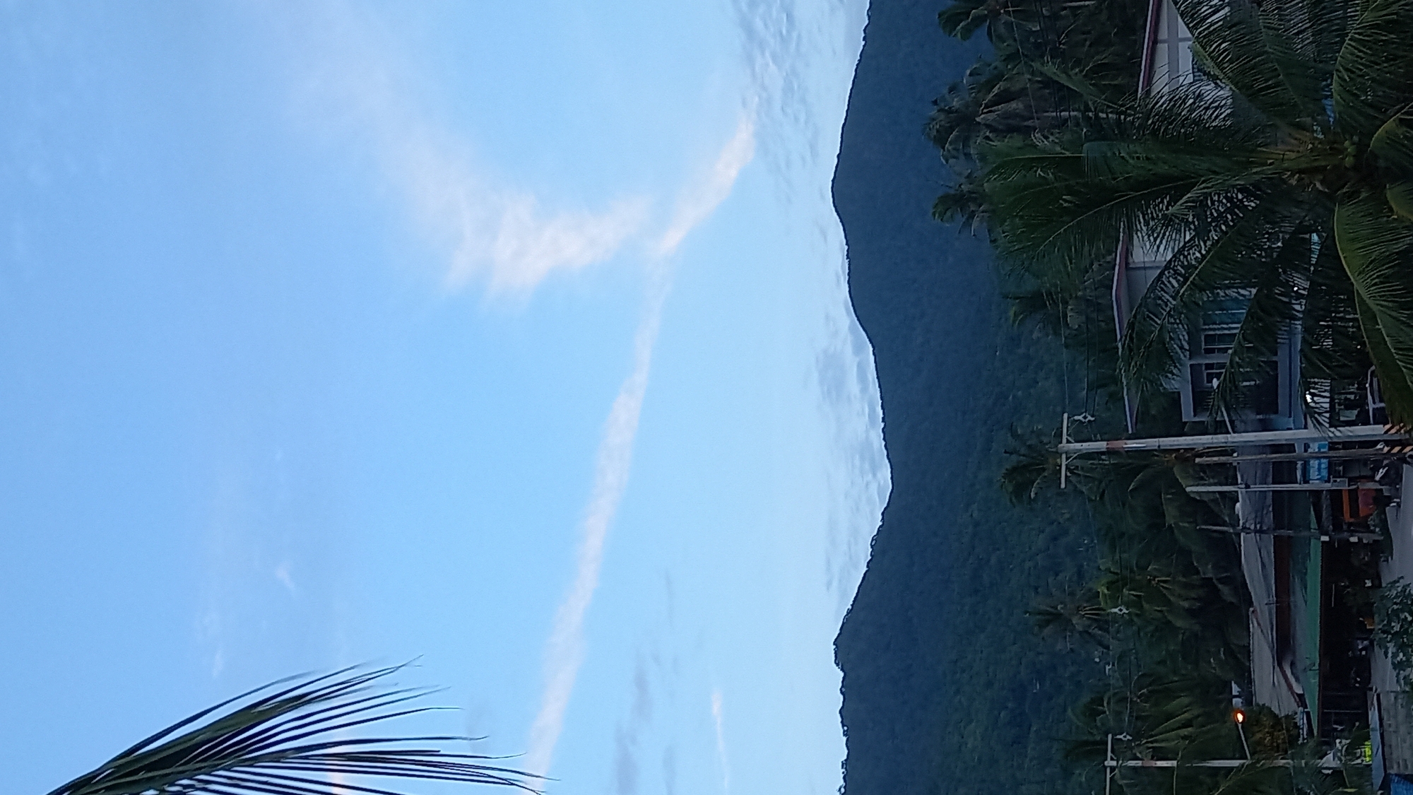 The image captures a serene, possibly twilight, view from Ko Pha-ngan District, Thailand, overlooking a coastal town nestled at the base of verdant mountains. The sky is a soft blue, painted with wispy white clouds, with a prominent streaky cloud formation resembling a "V" shape. In the foreground, a palm frond drapes in from the left, framing the scene. Below the mountains, a cluster of buildings, including what appears to be a modern two-story structure and smaller shops with awnings, are visible. Palm trees are scattered throughout the lower portion of the image, their fronds creating a textured canopy. Electrical poles with wires crisscross the town, indicating infrastructure. A motorcycle is parked on the left side of the road, and a few people are indistinctly present, suggesting a quiet, established community. The overall impression is of a peaceful, tropical locale.