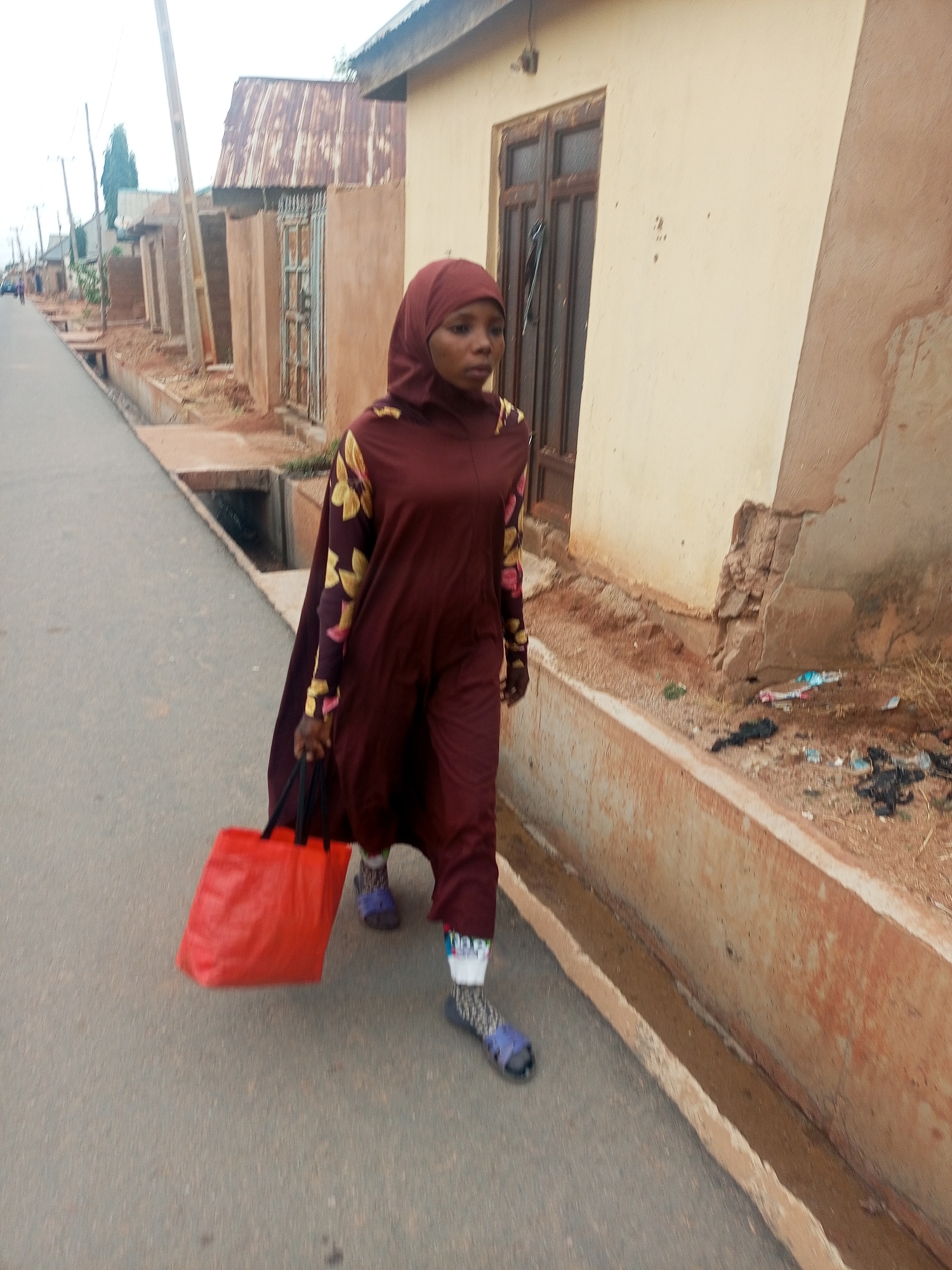 A woman is walking on a paved road in a residential area. She is wearing a maroon hijab and a matching long-sleeved dress with floral accents on the sleeves. She is carrying a red shopping bag. The road has drainage ditches on either side. Buildings are visible on the side of the road, some with corrugated metal roofs. The area appears to be a street in Tempure, Nigeria.