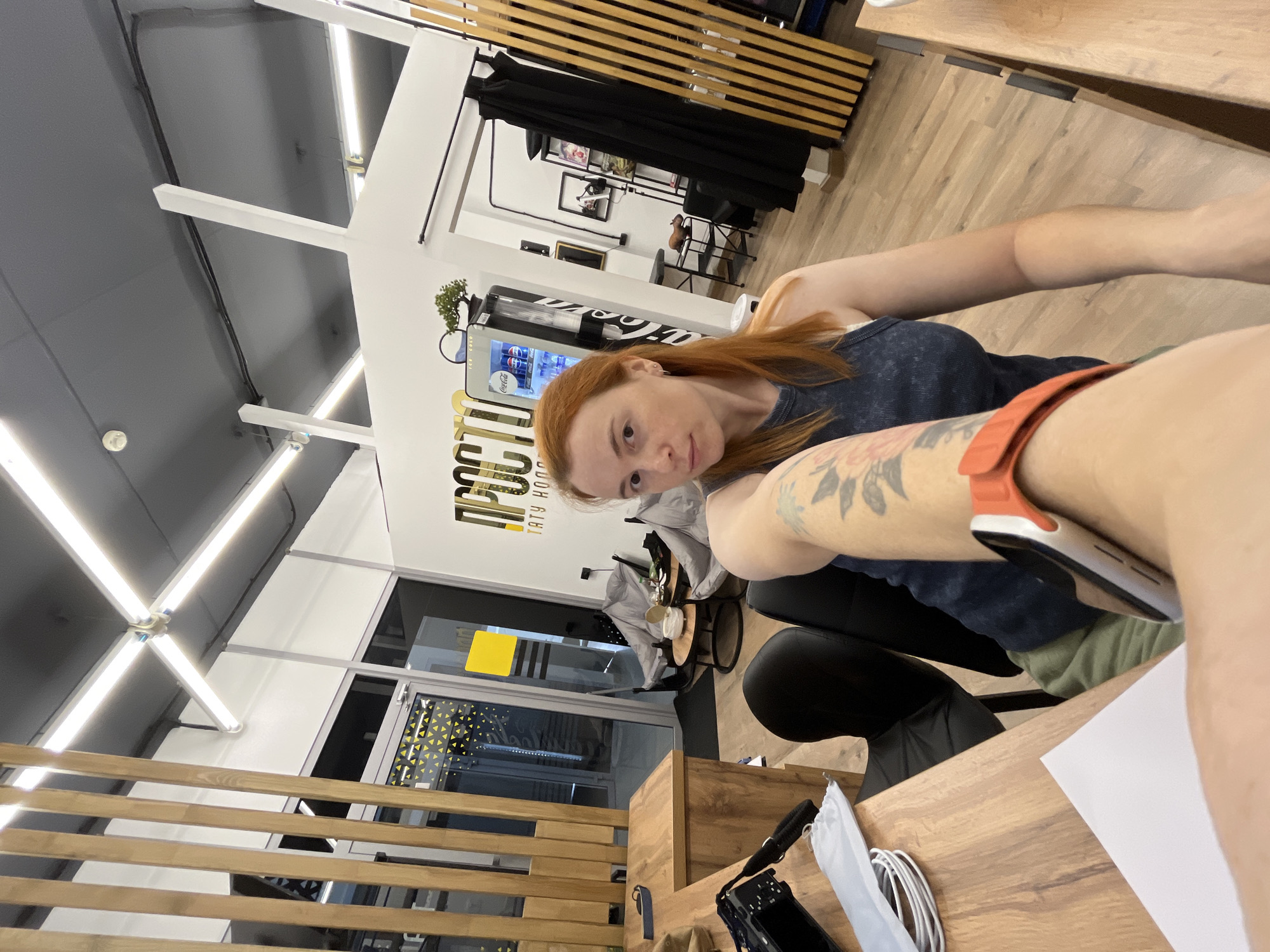 The image shows a woman with red hair, wearing a dark tank top and a green sweatshirt, sitting at a wooden desk in what appears to be a tattoo studio. She has tattoos on her left arm and is wearing an orange smartwatch. The studio has modern lighting with strip lights on the ceiling, wooden slat walls, and a glass door with a yellow sticker. In the background, there is a Coca-Cola vending machine, some framed pictures on the wall, and a black curtain. The overall environment suggests an urban setting, but without further context or identifiable landmarks, it is not possible to determine the specific location within Chaikovsky, Russia.