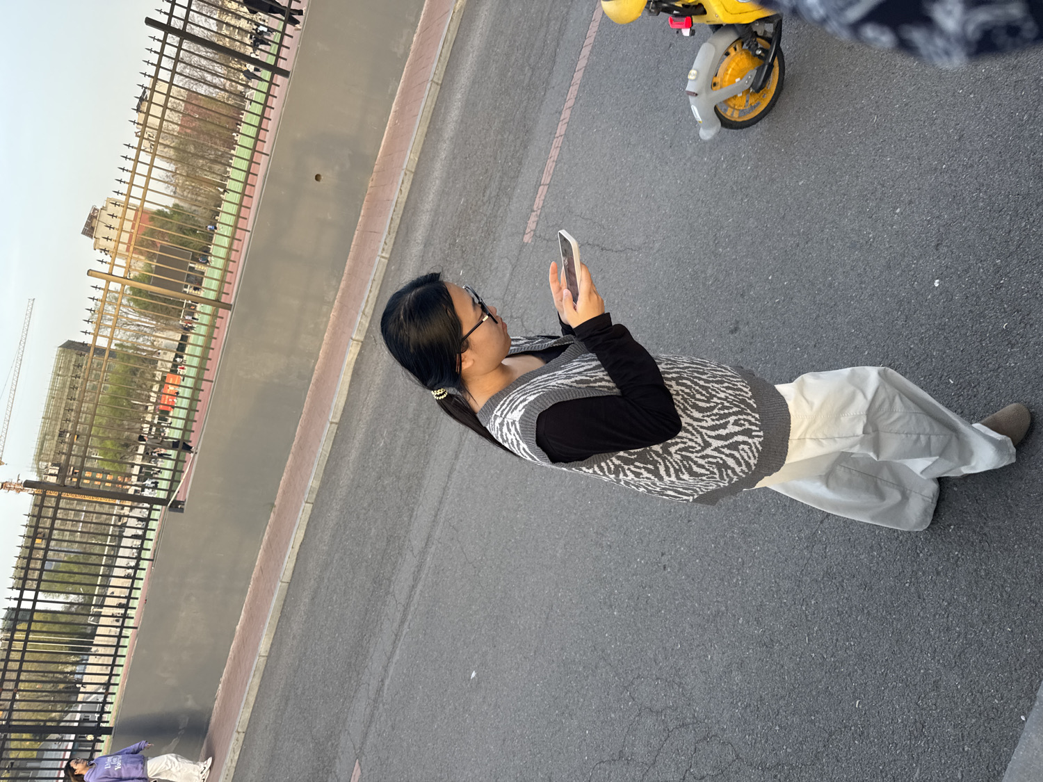 The image captures an outdoor scene featuring a woman engrossed with her smartphone, standing on an asphalt road. The woman has long dark hair pulled back with a scrunchie and wears glasses. Her attire consists of a black long-sleeved shirt underneath a grey and white zebra-print V-neck sweater vest, light-colored wide-leg pants, and light brown or grey slip-on shoes. She holds a white smartphone with both hands, directing her attention to its screen.
To the upper right, parts of a yellow shared bicycle, including its front wheel and handlebars, are visible. A black metal fence runs along the left side of the road, separating it from an area with green ground cover and sparse trees, possibly a sports field or park. In the background, under a bright, clear sky, multi-story buildings and a construction crane suggest an urban or developing setting. Another person in a purple hoodie is partially visible lying on the ground behind the fence in the lower-left corner.
The scene takes place during the daytime, likely late afternoon, with ample natural light. The ground is paved asphalt with some visible wear and a reddish-brown curb or strip bordering the fence line. The location context indicates this is in Zhongtun, China. No discernible text is visible on any objects or signage within the image.
