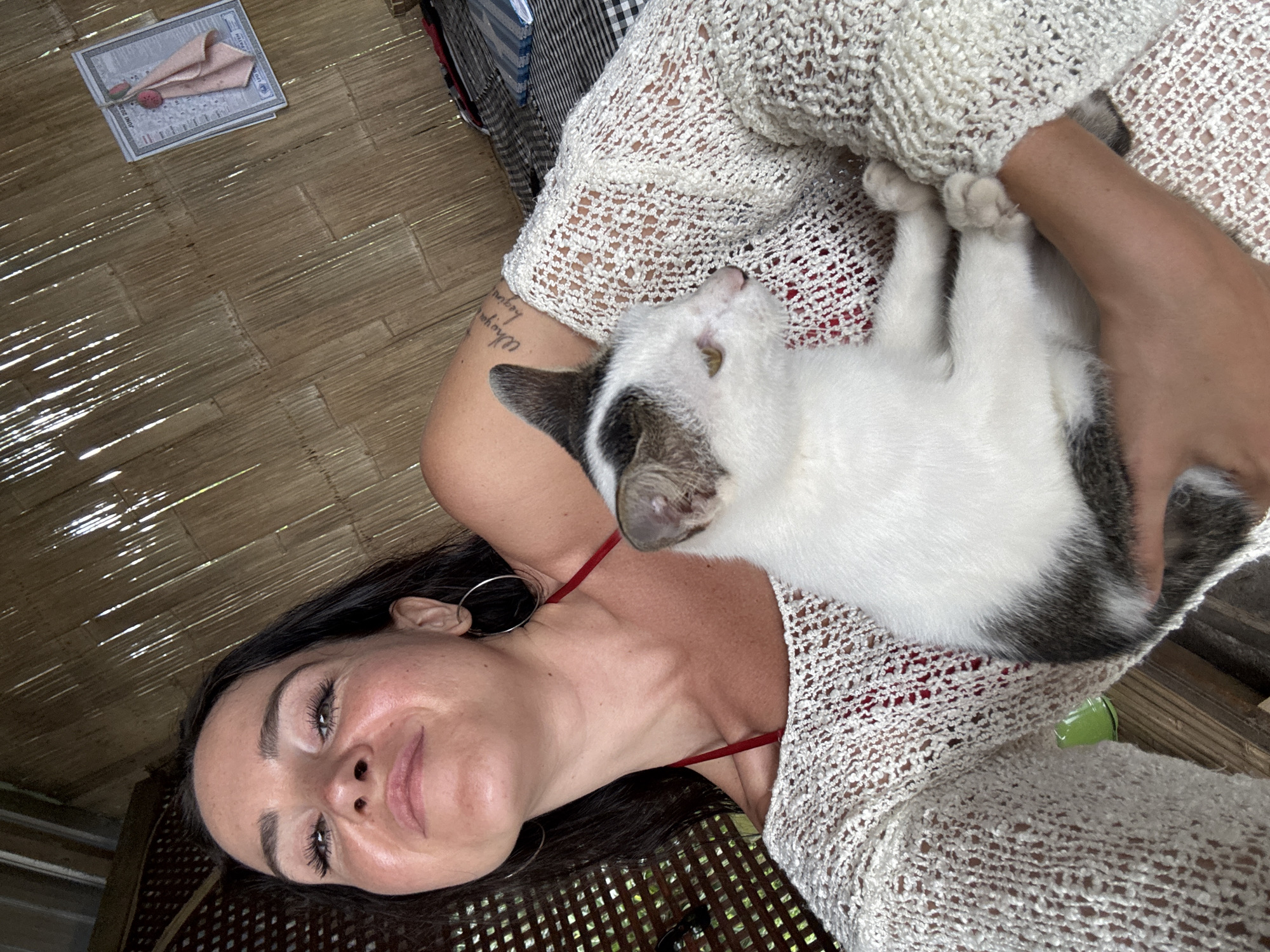 In an outdoor setting with a thatched bamboo wall and a wooden structure, a woman with dark hair and hoop earrings holds a white and gray cat. She is wearing a red bikini top and a loose-knit, off-white sweater. The cat is nestled in her arms, facing to the right. The scene appears to be a selfie, as indicated by the woman's pose and the camera's perspective. A small calendar and a pink cloth are visible on the wall behind her. The overall impression is one of casual relaxation.