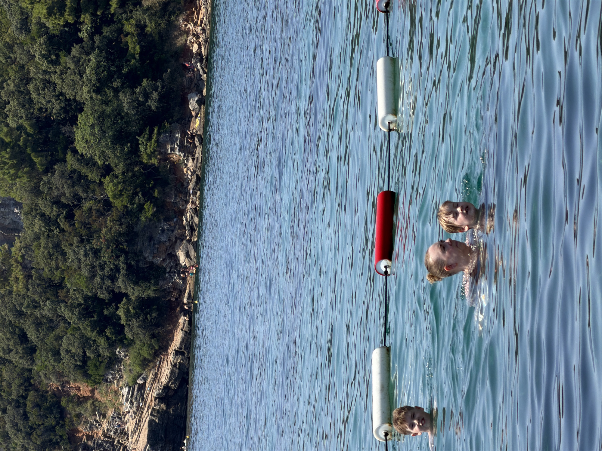 In the clear blue waters off the coast of Budva, Montenegro, a family enjoys a swim near a rocky, tree-covered shoreline. A woman with her hair tied up in a bun is swimming with two young boys. The boy on the left, with blond hair, is looking towards the camera. The boy on the right, also with blond hair, is looking away. In the background, a red and white buoy line stretches across the water. The shoreline is characterized by rugged rocks and lush green trees, with a few people visible sunbathing or walking along the rocky terrain. The water is calm with gentle ripples.