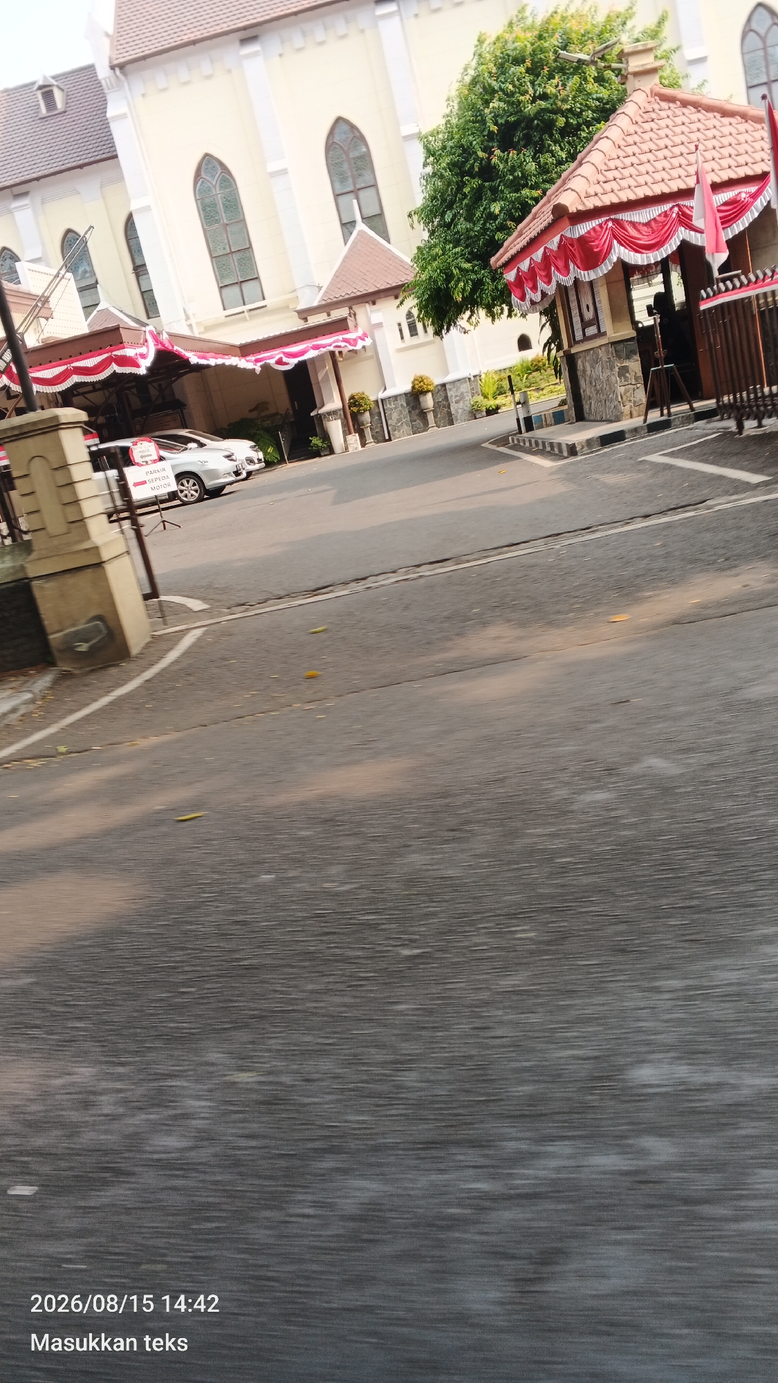 This is a photograph taken in the city of Malang, Indonesia, showing the exterior of a building that appears to be a church or a similar institutional building. The architecture is colonial-style, with a cream-colored facade and arched windows. In the foreground, a paved road is visible, with some scattered leaves. To the left of the road, a concrete pillar is present.
To the right of the building, a red and white striped awning is displayed over an entrance, with flags of Indonesia visible. Two cars are parked under another awning to the left of the building's main entrance. A sign on one of the cars reads "PARKIR SEPEDA MOTOR" which translates to "MOTORCYCLE PARKING."
The overall scene suggests a daytime setting, with sunlight casting shadows. The image has a timestamp at the bottom left corner indicating "2026/08/15 14:42" and the text "Masukkan teks".