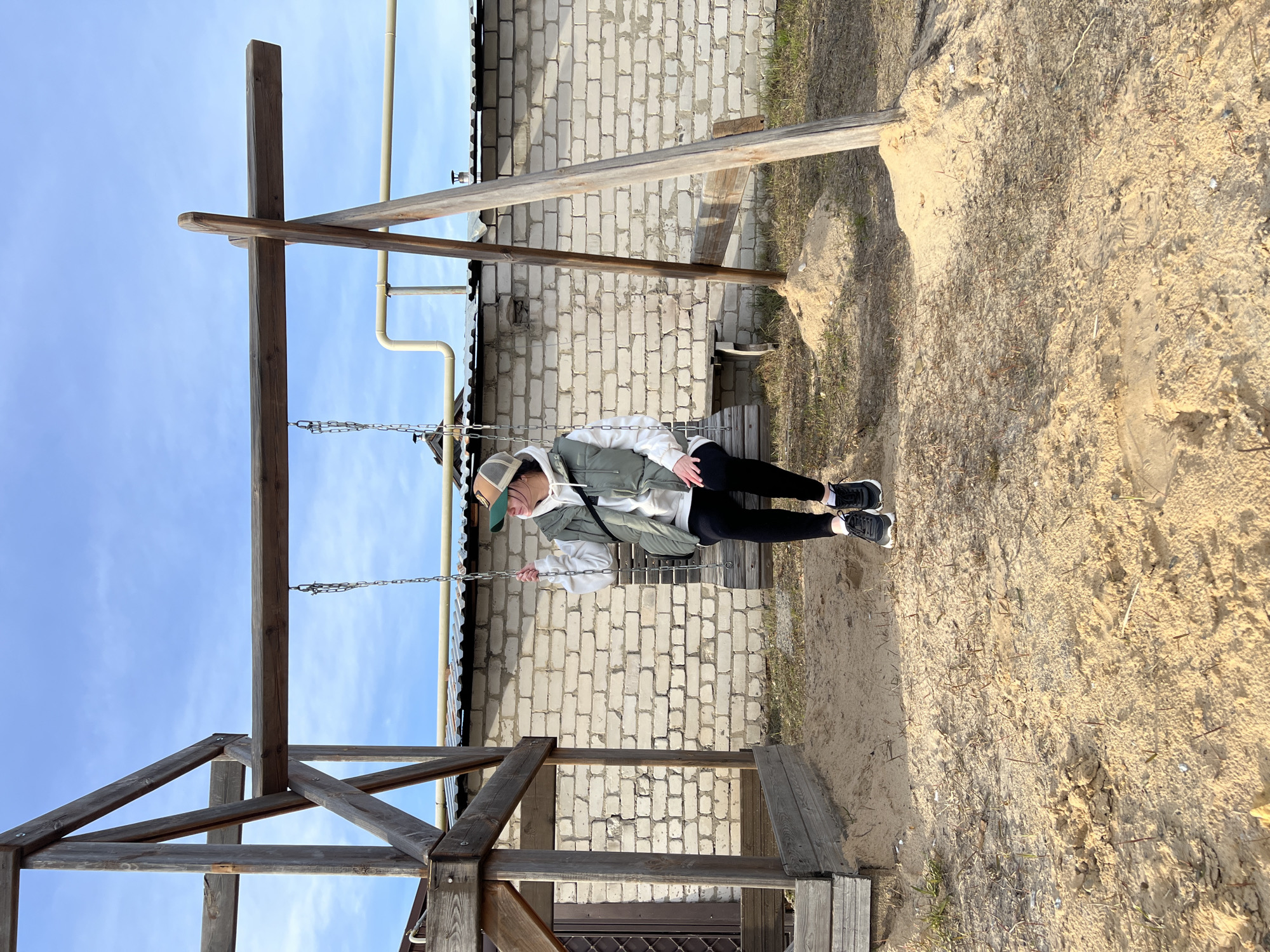 A young person wearing a cap, white hoodie, green vest, and black leggings sits on a wooden swing. The swing is part of a wooden structure. The ground is sandy. Behind the swing is a white brick wall. The sky is blue with light clouds. The location may be Kazan, Russia, but there is no explicit visual evidence to confirm this.