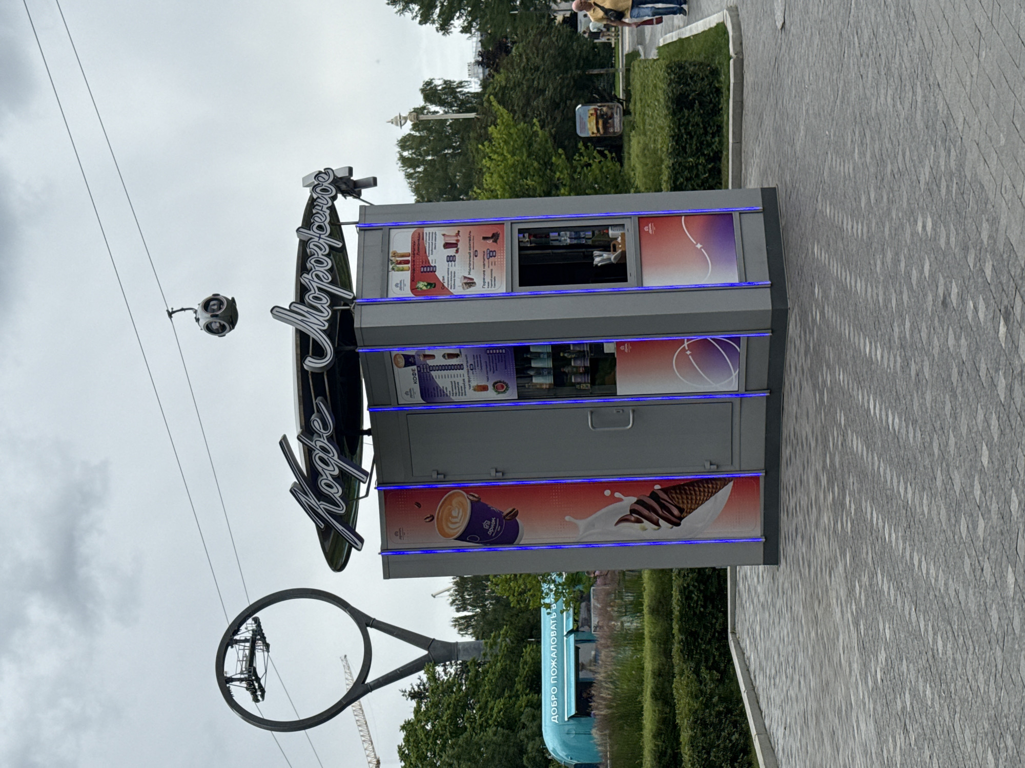 The image depicts an outdoor scene in Moscow, Russia, likely in a park or recreational area given the presence of trees, greenery, and a paved walkway. In the foreground, a modern, gray kiosk with bright blue LED lighting is prominently featured. The kiosk has large graphical advertisements on its sides, one displaying a coffee cup with latte art and a dripping ice cream cone, and another showing various beverage options and a menu. The text on the kiosk appears to be in Cyrillic script.
Above and behind the kiosk, a cable car is visible, suspended from wires that stretch across the frame. To the left, a large, circular metal structure with a suspended platform is also visible, possibly part of the cable car system or another attraction. In the mid-ground, a light blue food truck or kiosk with Cyrillic text that translates to "Welcome" is situated, surrounded by lush green bushes and grass.
In the background, several trees and some distant buildings are discernible under an overcast sky. On the right side of the frame, two individuals are walking away from the kiosk, on the paved walkway. One person is wearing a yellow shirt and blue jeans, and the other is wearing a dark shirt and blue jeans. The overall impression is a public urban space on a cloudy day, with a focus on a food and beverage service area connected to a cable car transportation system.