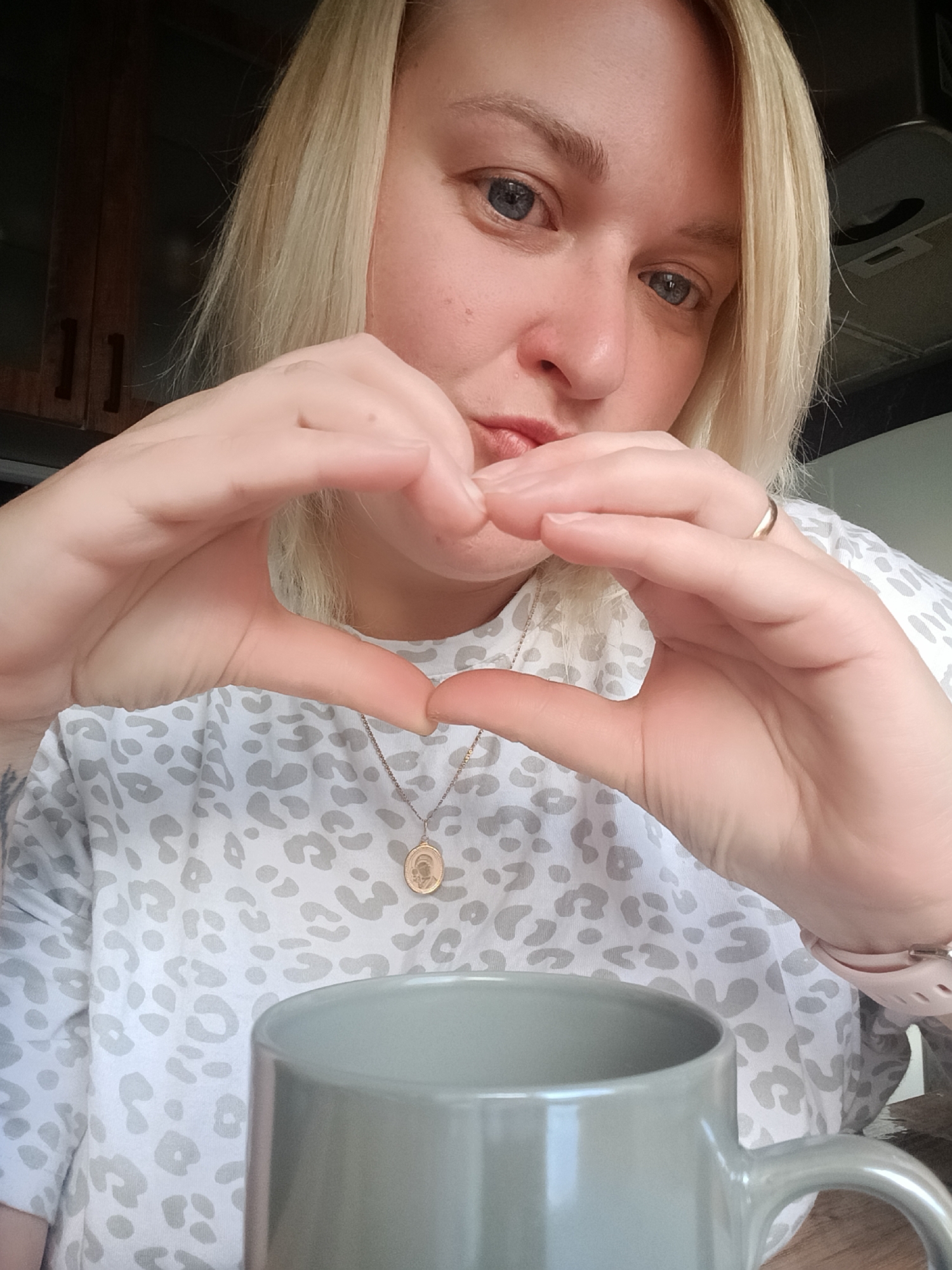 A fair-skinned, blonde-haired woman is posing for a selfie indoors. She is wearing a patterned, light-colored shirt and a gold necklace with a pendant. She is forming a heart shape with her hands near her face. A grey mug is visible in the foreground. In the background, dark wooden cabinets can be seen. The woman is wearing a wedding ring on her left hand and a pink wristwatch on her right. There is no explicit evidence in the image to confirm a location in the city of Chaikovsky, Russia.
