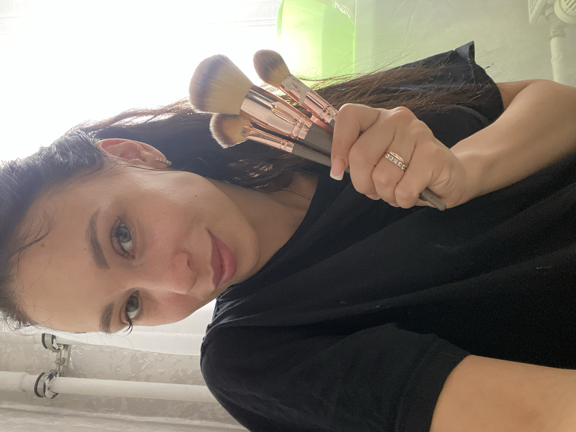 A female subject, estimated to be between 20 and 35 years old, is depicted in a medium-close shot, looking directly at the camera with a slight smile. She has dark hair, light eyes, and fair skin. The subject is wearing a black t-shirt and small hoop earrings on her left ear. On her left ring finger, she wears a gold-toned ring with multiple small stones. Her left hand, positioned centrally, holds four makeup brushes with dark handles, rose gold ferrules, and brown-blonde bristles; one brush exhibits white residue on its bristles.
The background is a brightly lit indoor environment. To the left, a white vertical pipe with silver clamps is visible against a light-colored, textured wall or curtain. To the right, a strong light source, likely a window, creates a high-key area. On a surface below this light source, a green plastic bowl containing a clear liquid is present, and part of a white radiator or heating unit is visible beneath the bowl. The setting appears to be a residential interior. The location in the city of Chaikovsky, Russia, cannot be determined from the visual evidence within the image.