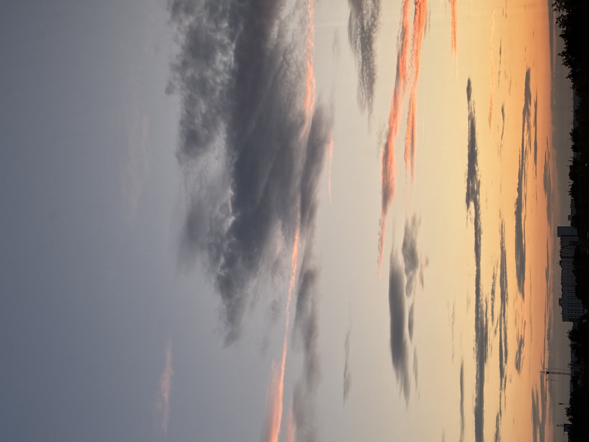 This is a vertical, low-angle shot of a sunset sky with scattered clouds. The sky transitions from a pale gray-blue on the left to a warm orange and yellow on the right. The clouds are a mix of dark gray and wispy white, with some catching the sunset light and appearing with pink or orange hues. Long, thin clouds streak across the sky, especially in the more colorful right side.
In the distance, along the right edge of the frame, there is a dark silhouette of trees and buildings. The buildings appear to be multi-story residential structures. A construction crane is visible between the trees and buildings, suggesting an urban or developing area.
The time of day is clearly sunset due to the colors in the sky and the lighting conditions. The weather appears calm, with the clouds creating visual interest but not indicating any severe conditions. There are no people or animals visible in the frame. The overall mood is serene and picturesque, highlighting the beauty of the natural light display. No text is visible in the image.