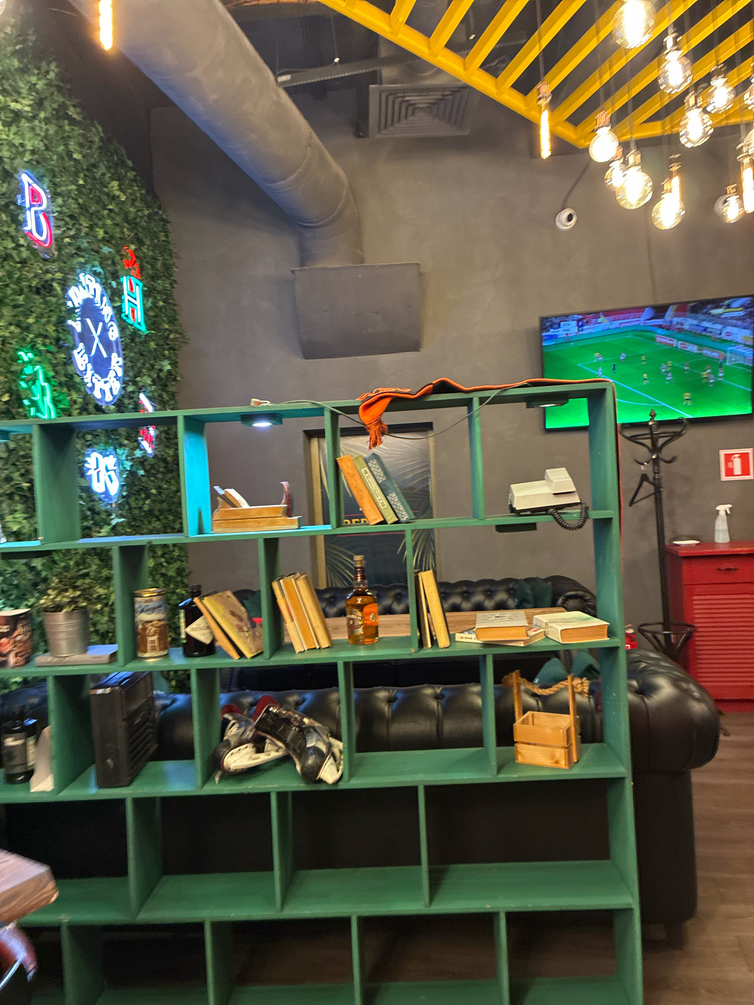 The image depicts an interior scene, possibly a bar or lounge, with a dark grey wall and a large green shelving unit. The shelving unit contains various items, including books, bottles of alcohol, a retro telephone, and decorative objects. A large television screen on the wall shows a soccer game. The room is decorated with neon signs on a wall covered in artificial greenery. The ceiling has exposed yellow beams and decorative light bulbs. A red cabinet and a coat rack are visible in the background. The scene is well-lit, with a mix of ambient and artificial lighting. There are no people visible in the image. The overall atmosphere suggests a modern, casual establishment.