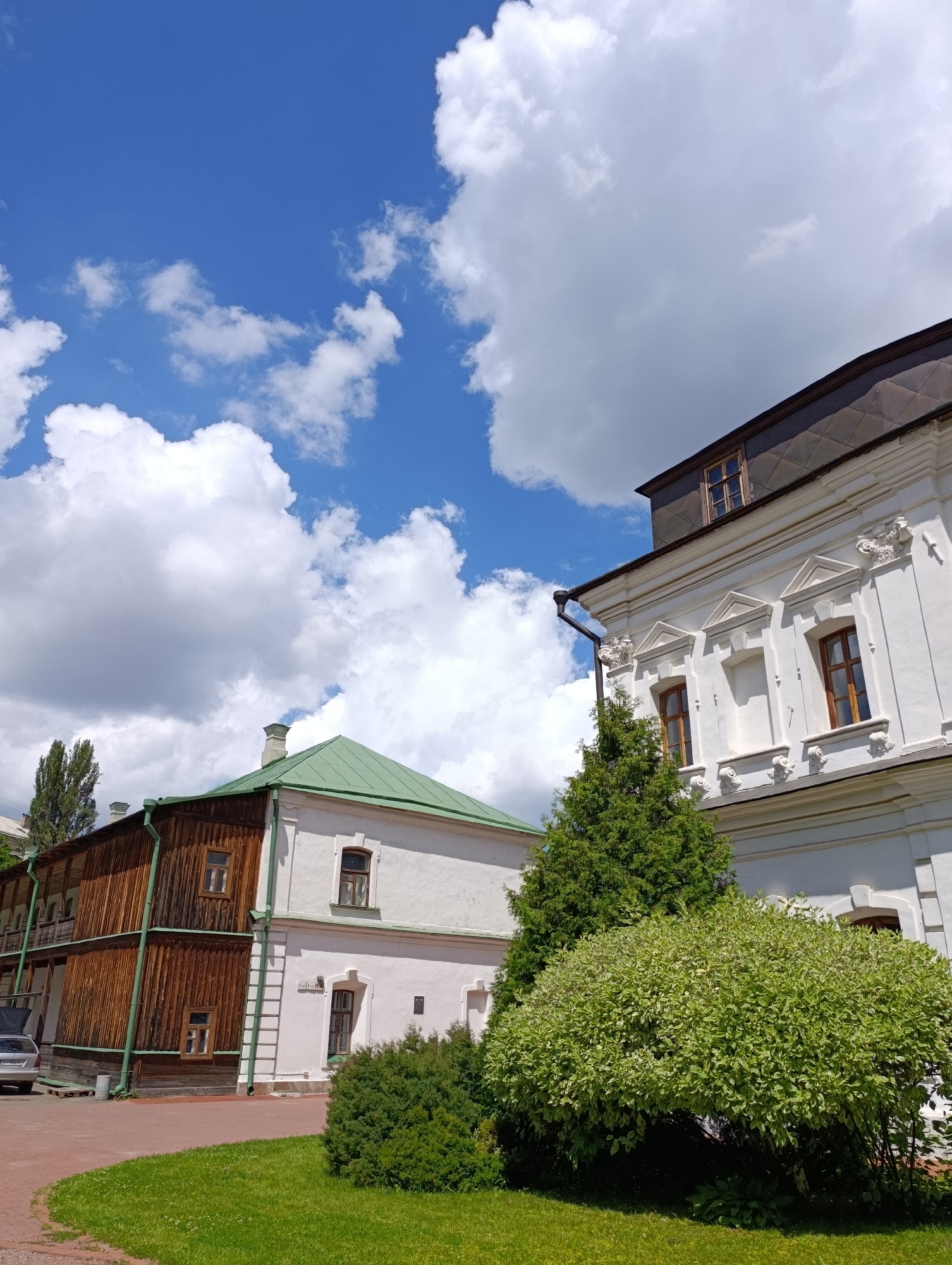 The image depicts an outdoor scene with architectural elements and natural scenery. In the foreground, a well-maintained lawn is visible, with a paved walkway curving through it. Adjacent to the lawn are several buildings. To the left, a wooden building with multiple stories features a weathered wood facade and a green gutter system. It has several windows and a covered balcony area. To the right, a white plastered building with classical architectural details is prominent. This building has multiple windows, decorative cornices, and a dark, patterned roof.
In front of the white building, a lush, green shrub is in full bloom, its leaves a mix of green and white. Behind this shrub, a tall, slender evergreen tree stands. Further in the background, a large expanse of blue sky with scattered white cumulus clouds dominates the upper portion of the image, suggesting fair weather. The lighting indicates it is daytime, likely mid-morning to early afternoon, due to the bright sunlight casting shadows.
The scene appears to be a historical or cultural site given the architectural styles of the buildings. There are no people visible in the image. There is no discernible text within the frame. The overall impression is one of a serene, well-preserved historical area bathed in pleasant daylight.