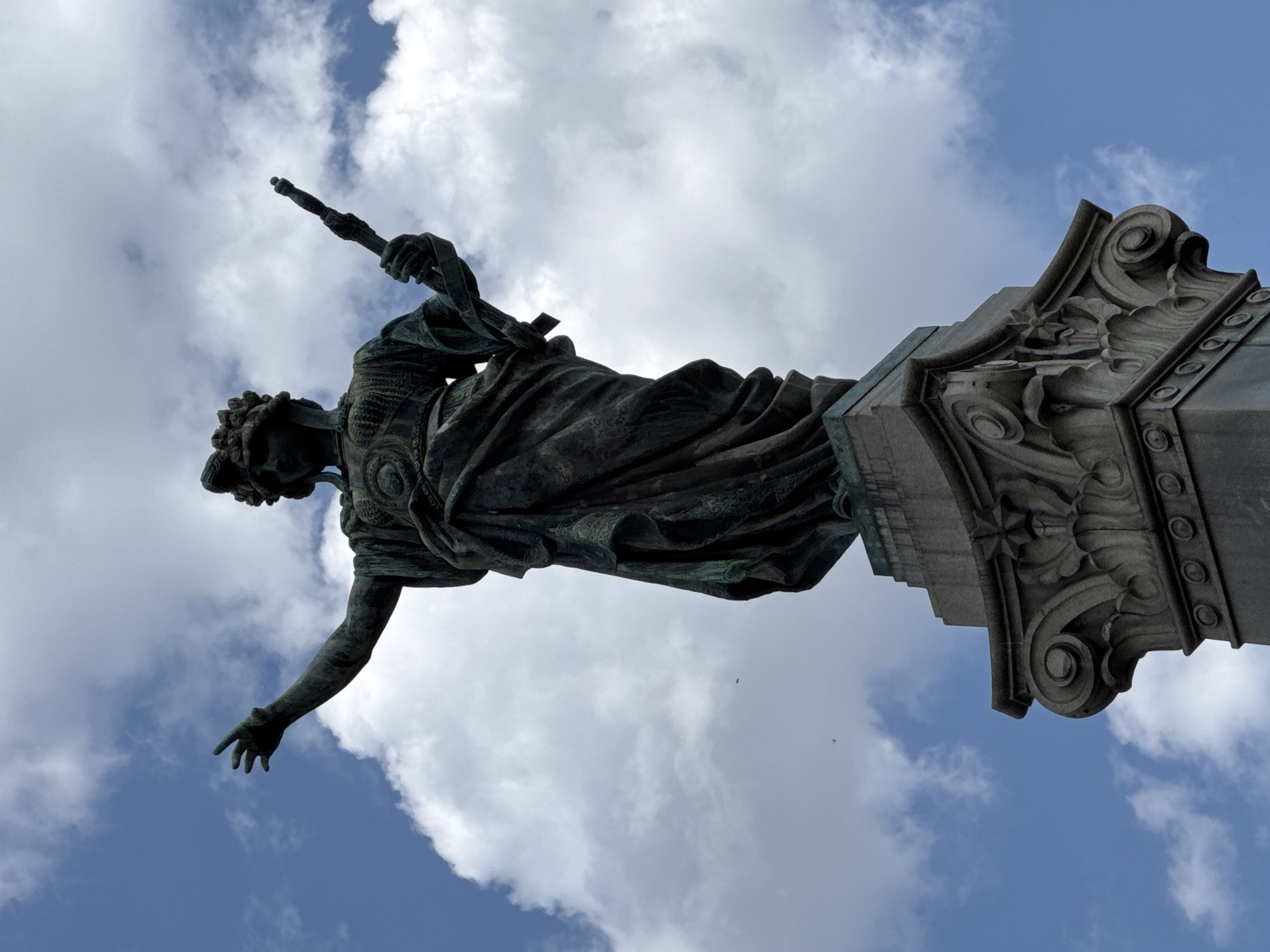 This is a low-angle, sideways photograph of a weathered bronze statue of a robed figure, possibly allegorical, holding a staff or scepter. The statue appears to be very old and is mounted on a stone pedestal with intricate carvings. The figure is depicted with its left arm outstretched, as if reaching or pointing. The background is a bright blue sky filled with fluffy white clouds, suggesting a daytime scene. The lighting indicates it is likely midday or early afternoon. The clouds are prominent, with some casting soft shadows on the statue and pedestal. The perspective is from below, looking up at the statue, emphasizing its height and grandeur against the sky. The overall mood is solemn and majestic. There is no visible text in the image. The scene is set outdoors, with the sky serving as the primary backdrop. The statue's details, such as the fabric folds of its robe and the facial features, are softened by time and the elements. The ornate carvings on the pedestal suggest a historical or monumental monument.