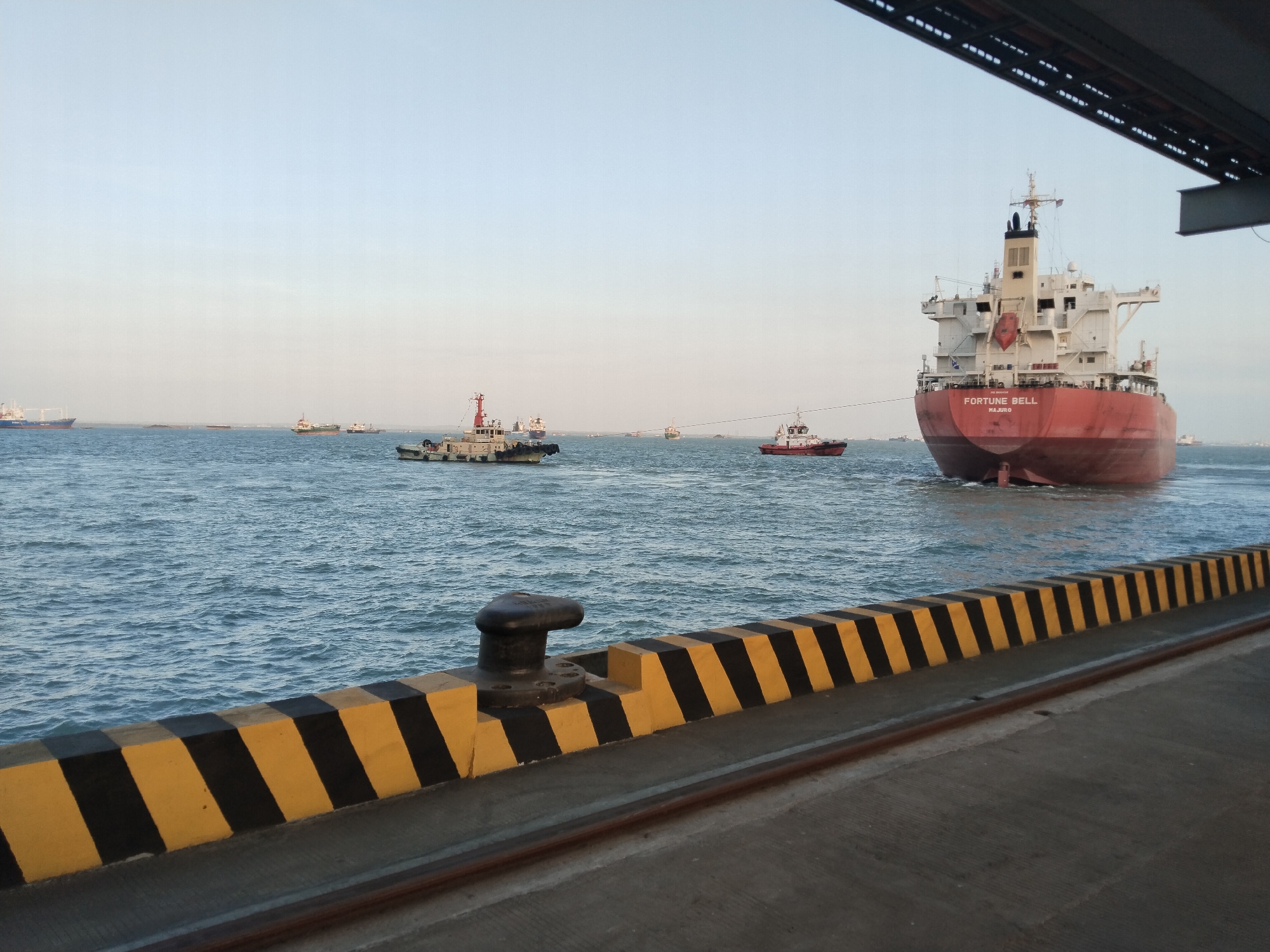 The image depicts a harbor scene at sunset. In the foreground, a concrete pier is visible, marked with alternating yellow and black diagonal stripes along its edge, with a bollard on the left. The sea is calm, with moderate waves. Several ships are present in the water. On the right side, a large, red-hulled cargo ship named "FORTUNE BELL" is partially visible, with a tugboat attached to its stern, seemingly assisting it. In the middle ground, a smaller tugboat is also visible, connected by a tow line to another vessel. More ships, including some smaller vessels, are scattered across the horizon. The sky is clear and light blue, transitioning to a lighter hue at the top, indicating the time of day is either early morning or late afternoon. The overall atmosphere is one of maritime activity. The scene is located in the port of Pernajuh, Indonesia.