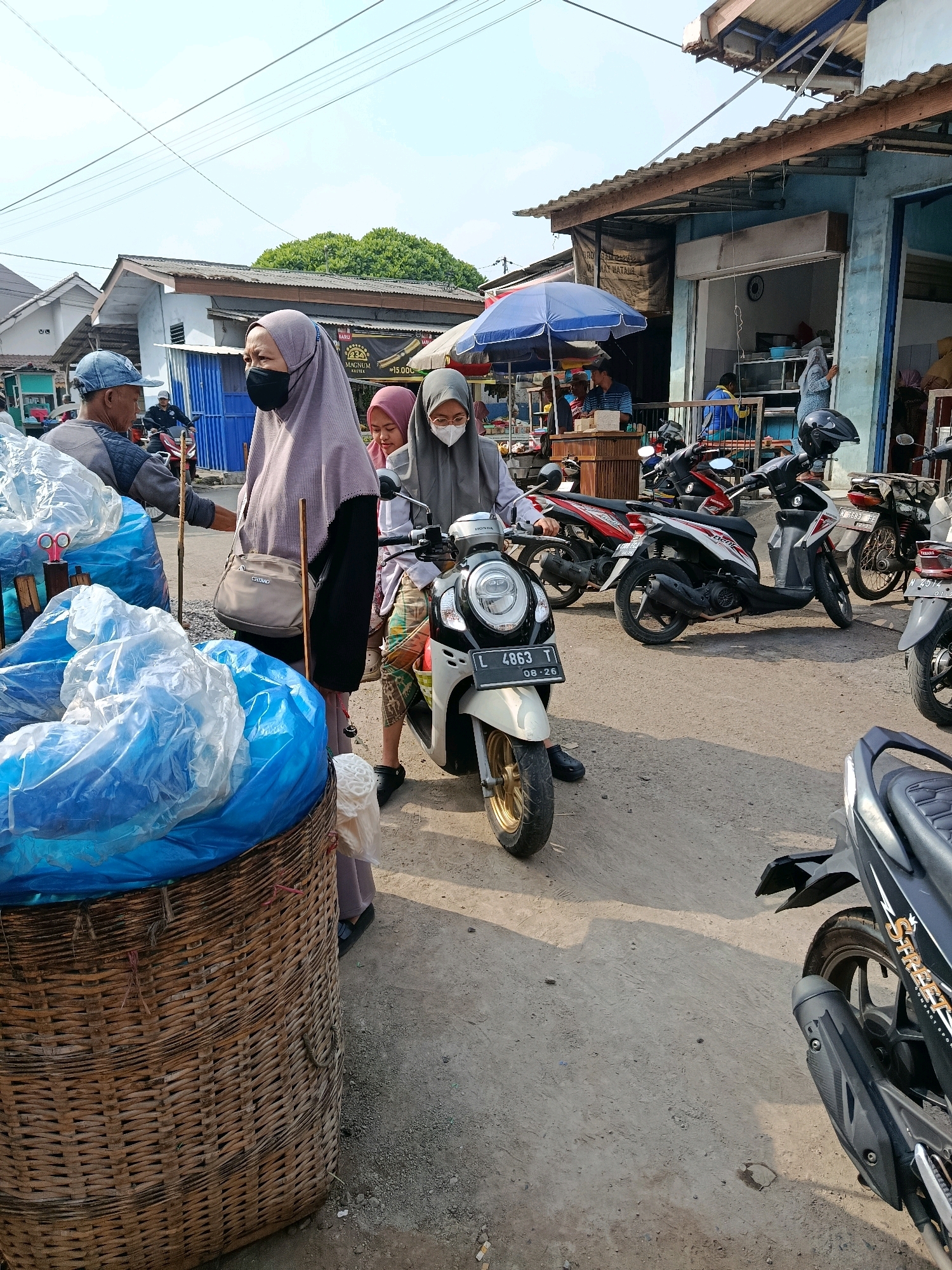 The image depicts a street scene in Bululawang, Indonesia, with several people and motorbikes. In the foreground, a large woven basket is filled with blue plastic-wrapped items. To the right of the basket, a woman wearing a hijab and a face mask is standing next to a motorbike, on which another woman with a face mask and glasses is seated. A man wearing a cap is to the left of the basket, facing the woman in the hijab. In the background, several motorbikes are parked. Buildings with awnings line the street, suggesting a market or commercial area. The ground is unpaved, with some gravel and dirt. The lighting indicates it is daytime, with shadows cast by the structures and vehicles.