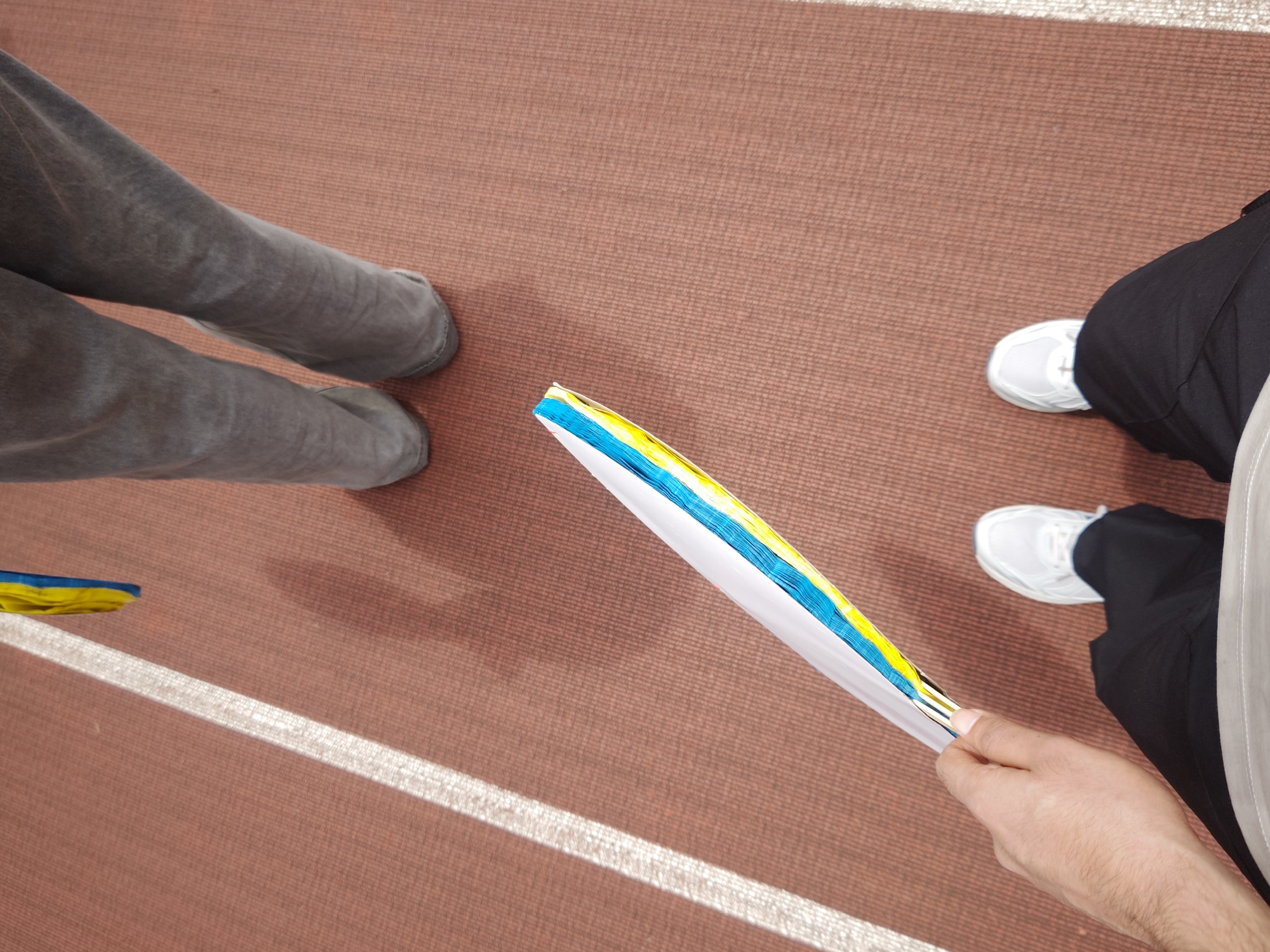 The image, taken from a high-angle, downward perspective, shows the lower bodies of two individuals standing on a reddish-brown textured surface resembling an indoor track or athletic hall. A distinct white line runs horizontally across the ground, dividing the frame.
On the left side of the frame, one person's legs are visible, clad in grey, possibly denim or soft fabric, pants, ending in grey, low-profile shoes or slippers. To the right, another person wears black pants and white athletic sneakers. This individual is holding a long, thin, fan-like object with their right hand, which also shows a white sleeve. The object features prominent bright blue and yellow colors along its edges, reminiscent of the Ukrainian flag.
The setting appears to be an indoor environment due to the consistent texture of the ground and the clear definition of the white line, commonly found in sports facilities. The bright lighting suggests either strong overhead artificial lights or natural light from a large window. The two individuals are seemingly standing in close proximity, possibly engaged in conversation or waiting. The location context provided indicates this scene is in the Chongqing High-tech Zone, China. No visible text is present in the image.
