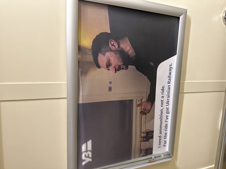 This image displays a vertically oriented, framed poster mounted on a plain, light-colored wall, characteristic of an interior space such as a train carriage. The poster features a photograph of Volodymyr Zelenskyy, President of Ukraine, shown from the chest up in profile, facing left. He has short dark hair and a beard, and wears a dark long-sleeved shirt with an embroidered emblem resembling the Ukrainian trident on the left chest. His right hand is raised in a speaking or gesturing posture. The background of the photograph within the poster includes elements suggesting an interior compartment, such as a window frame and wall details, with the numbers "16 46" visible on a panel. The "УЗ" (Ukrainian Railways) logo is present in the upper left corner of the poster's image. Below the photograph, white text states: "I need ammunition, not a ride. For the ride I've got Ukrainian Railways." The poster itself is enclosed in a silver-colored metallic frame. A metallic fixture is partially visible at the bottom edge of the overall image. The specific location within the city of Kvitneve, Ukraine, cannot be determined from the visual information provided.