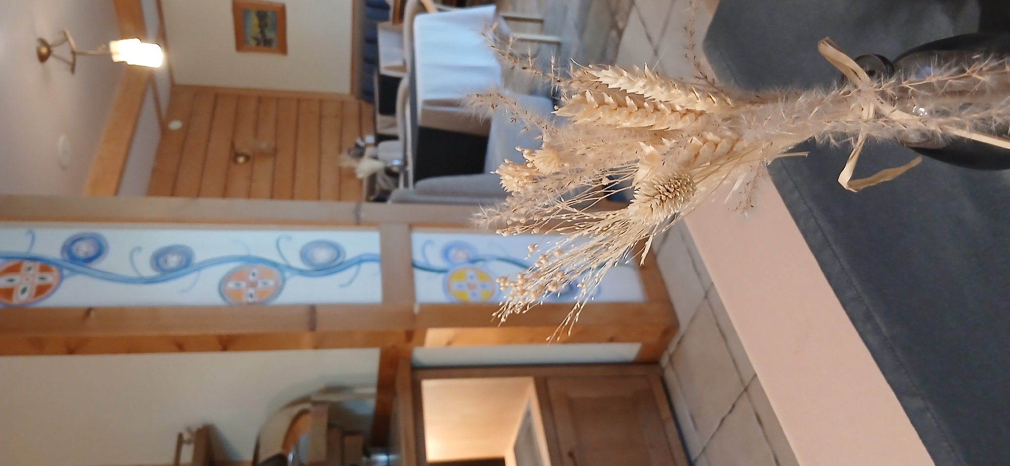 The image depicts an indoor scene, likely a restaurant or cafe, with a focus on a vase of dried decorative grasses in the foreground. The dried grasses are light tan and feathery, arranged in a dark, possibly glass, vase. Behind the vase, a table with a light pink runner is visible. Further in the background, there is a wooden pillar with decorative tiles featuring circular patterns in orange and blue. The environment appears to be made of wood and light-colored walls. In the mid-ground, tables are set with white and dark tablecloths, and chairs are visible. The ceiling has exposed wooden beams and a hanging light fixture. A framed picture hangs on a wooden-paneled wall in the background. The overall impression is a rustic and cozy interior setting. No people are visible in the image. The location is unknown.