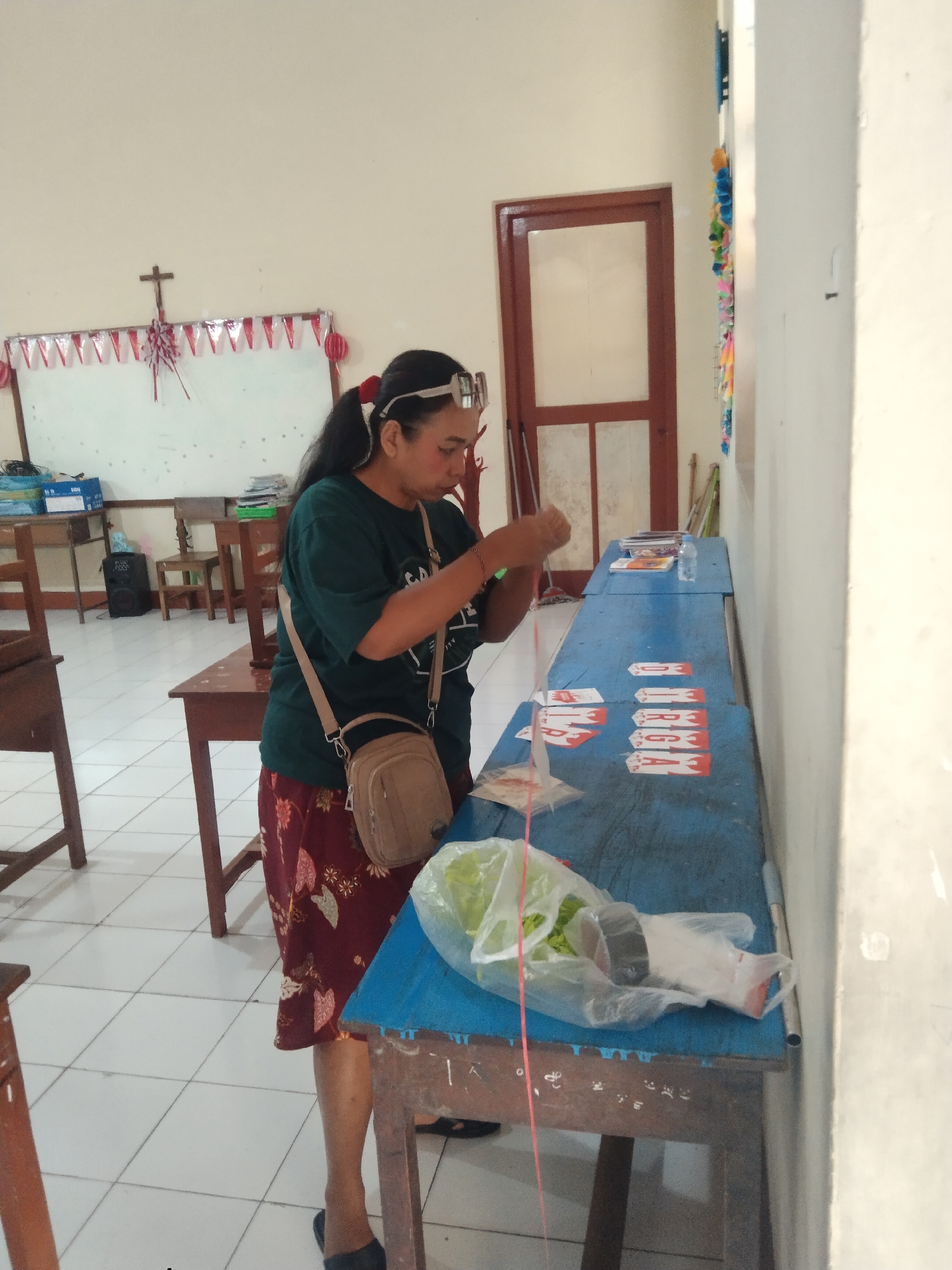 A woman with dark hair tied back in a ponytail and wearing a dark green t-shirt and a patterned red skirt is standing at a long, blue table, engaged in an activity. She has a beige crossbody bag over her shoulder and wears glasses on her forehead. Her right hand is holding a string or thin material, and her left hand is extended towards the table. On the table, there are several white strips with red markings, arranged in rows. Some of these strips have red rectangular shapes and white lettering. In front of the woman, on the table, is a clear plastic bag containing green vegetables. The room has a tiled floor and cream-colored walls. A whiteboard decorated with red and white pennant banners and a cross is visible on the wall in the background. Several wooden tables and chairs are also present in the room. A dark wooden door with glass panes is seen in the background. The overall scene suggests an indoor setting, possibly a classroom or community hall.