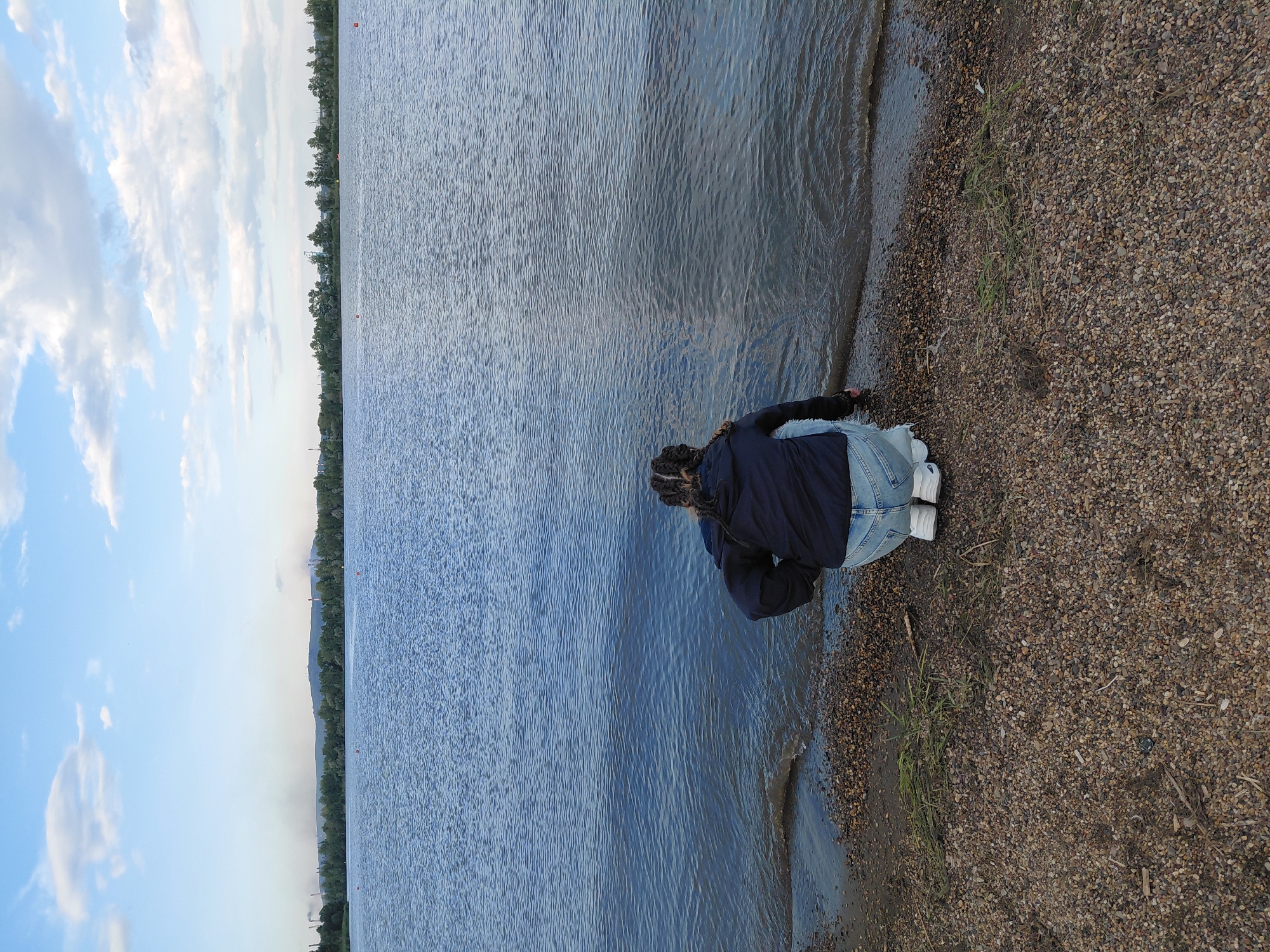 A young person with braided hair is crouching on a pebbly shore next to a large body of water. They are wearing a dark hooded jacket and light-colored denim shorts. The water is calm with small ripples, and there are buoys visible in the distance on the surface. The background shows a treeline and a hilly landscape with industrial structures and faint smoke plumes, indicative of Magnitogorsk, Russia. The sky is partly cloudy with a soft, diffused light.