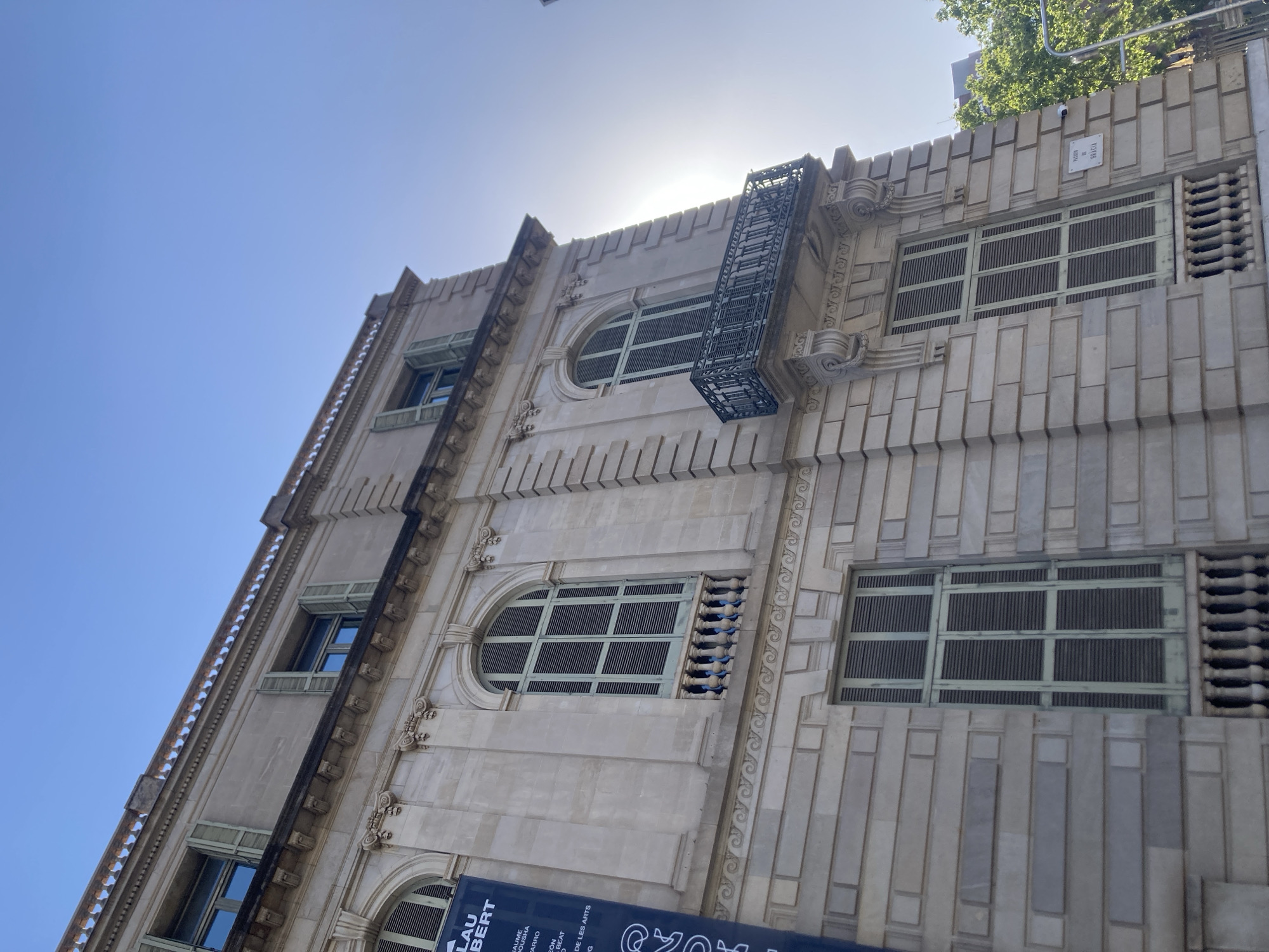 This image captures a low-angle view of an ornate building facade against a bright, clear sky, suggesting a sunny day. The architecture features a light-colored stone or concrete material with distinct vertical and horizontal layering, giving it a textured appearance. Several windows are visible, many with green slatted coverings. Some windows are arched, while others are rectangular. A decorative black metal grate, resembling a small balcony or fire escape, protrudes from the building.
A sign on the building reads "PASSES DE GRACIA," indicating the location is likely Passeig de Gràcia in Barcelona, Spain, a well-known boulevard famous for its modernist architecture. Another visible sign, partially obscured, includes names like "LAU BERT," "JAUME," "BOUSHA," and "ARRO," which might be related to an exhibition, event, or the building's occupants. The presence of a small security camera mounted on the facade is also noticeable. The sun is high in the sky, creating a slight lens flare and casting a bright light on the upper parts of the building, with some trees visible at the very top. The overall impression is one of architectural grandeur on a bright, clear day in a bustling urban environment.