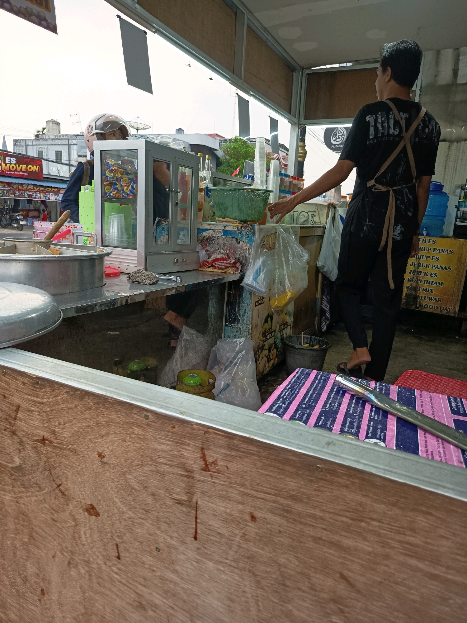 The image depicts an outdoor street food stall in Banjarmasin, Indonesia. Two individuals are present. In the foreground, a person wearing a helmet is partially visible behind a metal counter, possibly operating the stall. In the midground, a person wearing a black t-shirt and an apron stands facing away from the camera, tending to the stall. The stall is equipped with various items including a glass display case with packaged goods, a large metal pot, plastic cups, a green basket, and a blue water dispenser. Behind the stall, a yellow sign displays handwritten text in Indonesian, listing food and drink items such as "ES", "JERUK PANAS", and "KOPI HITAM". The environment appears to be a busy urban setting, with buildings and a street visible in the background. The overall scene suggests a typical day of business at a local food vendor.