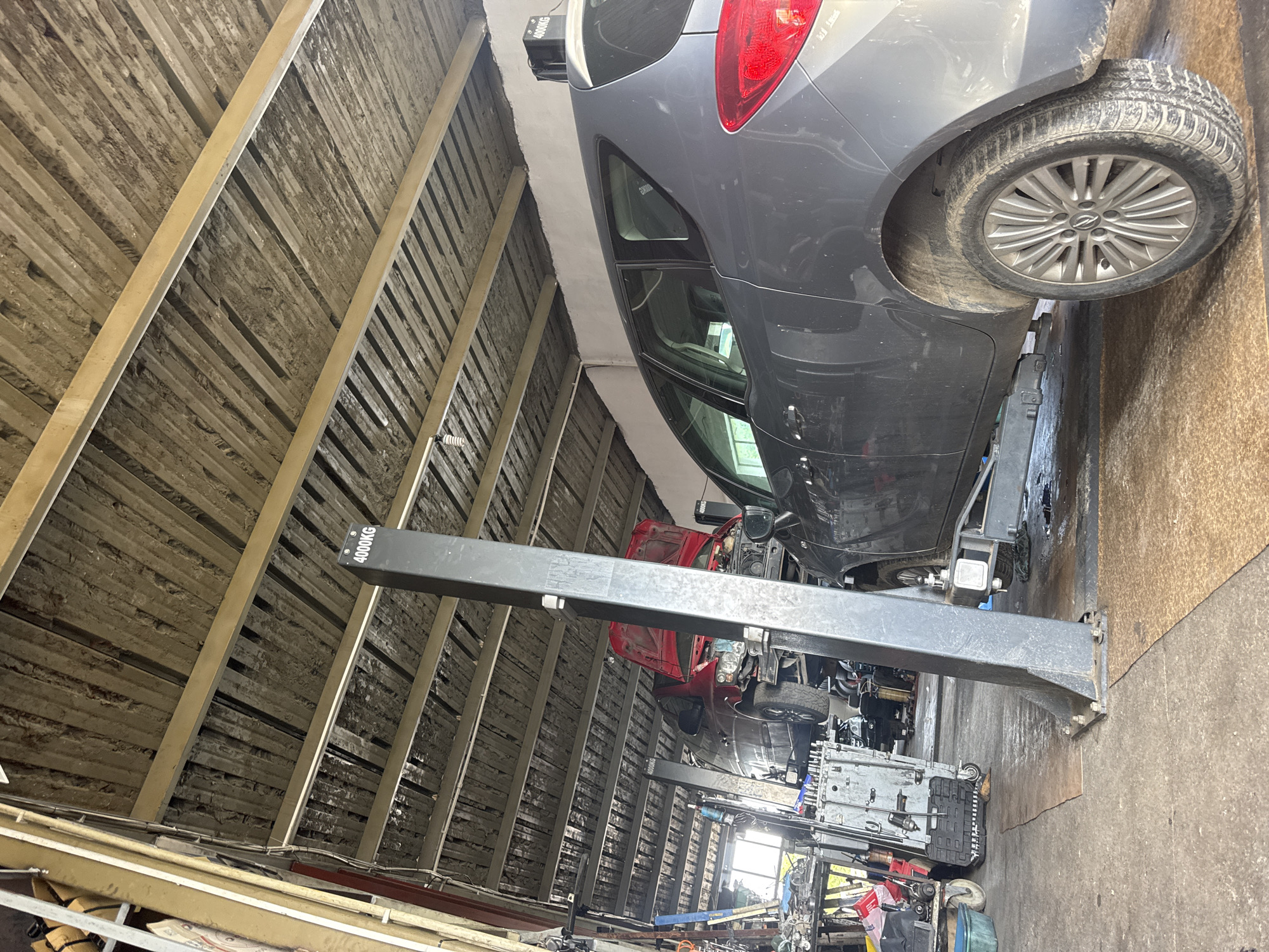 The image shows the interior of a garage, potentially located in Yambol, Bulgaria, though this is not definitively confirmed. A grey hatchback car is elevated on a four-post vehicle lift. A red car is visible underneath the grey car on a separate lift. Various tools and equipment are scattered around the garage floor. The ceiling is unfinished, revealing wooden beams and insulation material.